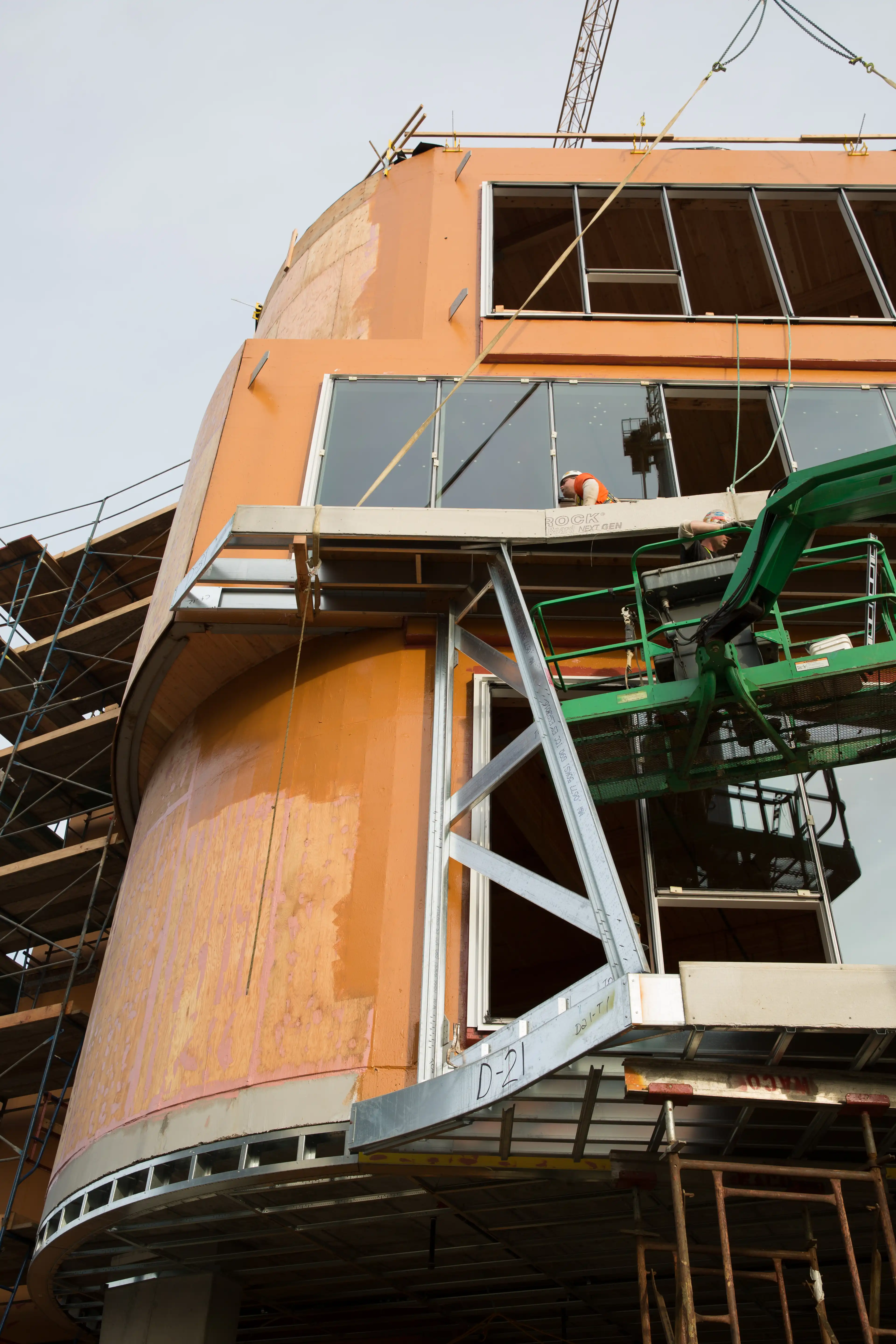 Construction crew works along a curved façade with large glass openings and orange sheathing, using lifts and scaffolding to install framing for Passive House apertures.