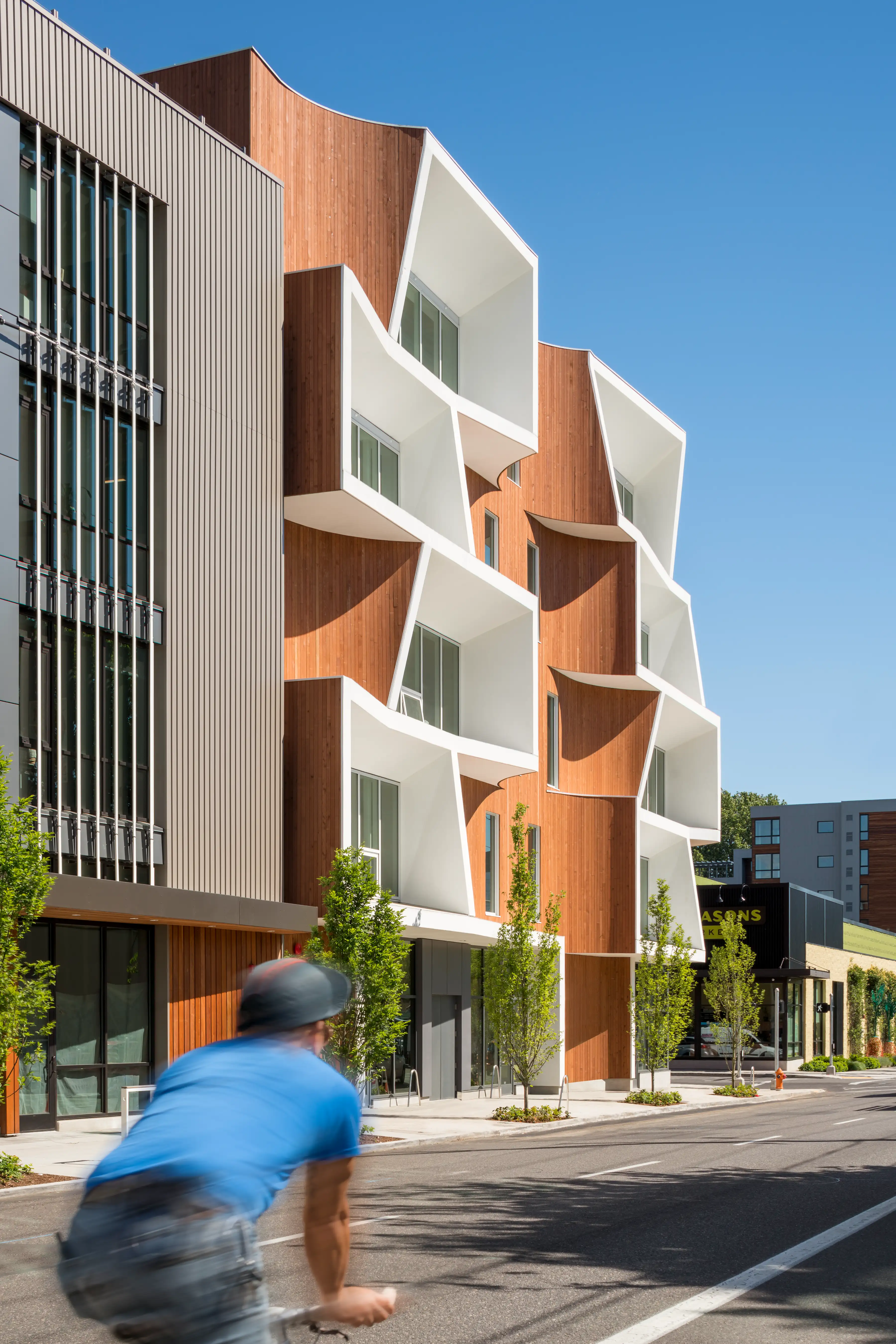 Modern building with wood cladding and sculptural white apertures, with a cyclist passing along the tree‑lined sidewalk.