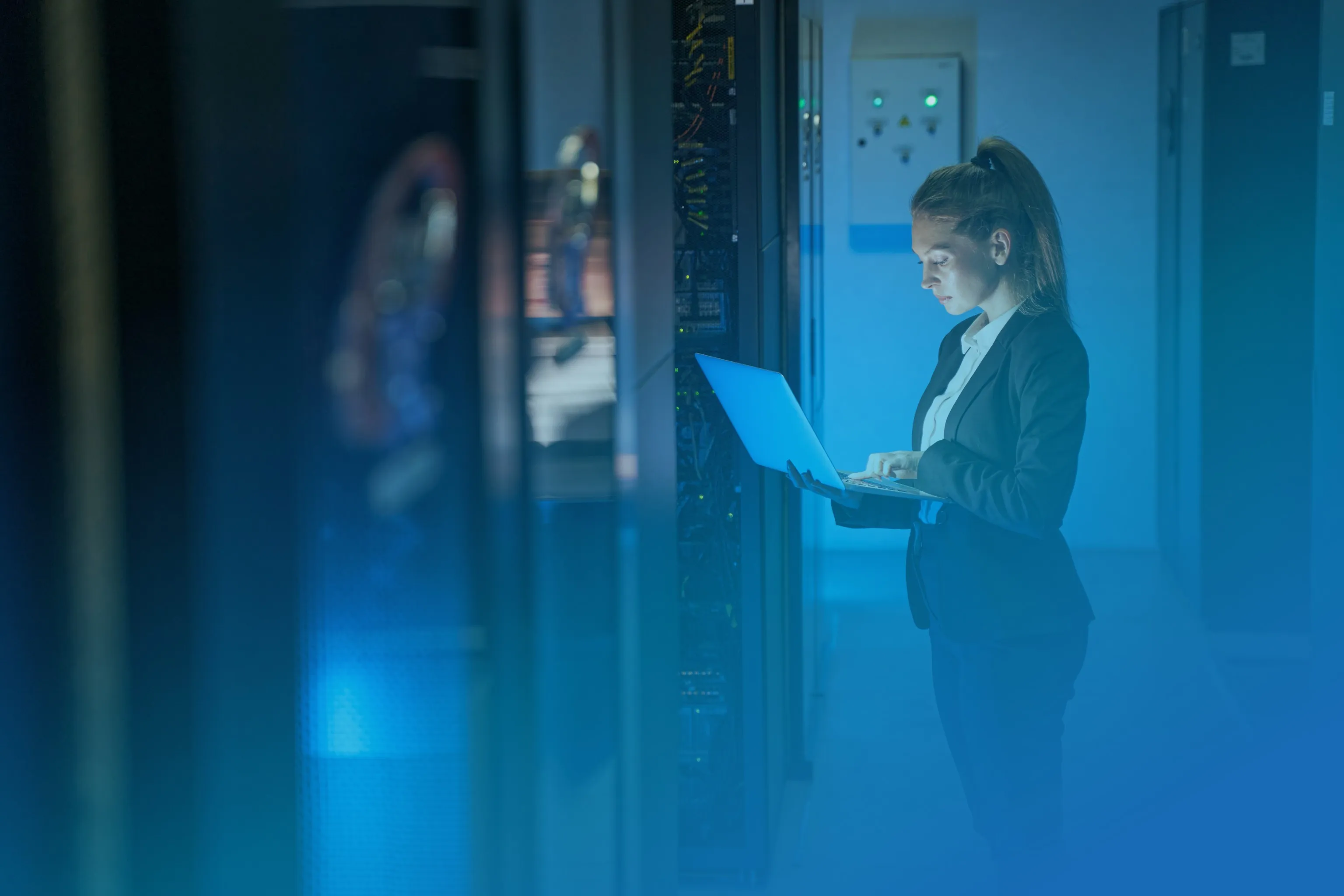 Woman in business attire working on a laptop inside a server room with network equipment.