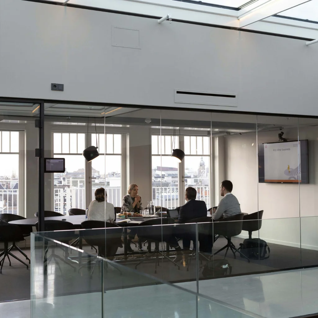 Four people sitting around a table in a glass-walled meeting room with windows overlooking a cityscape during daytime.