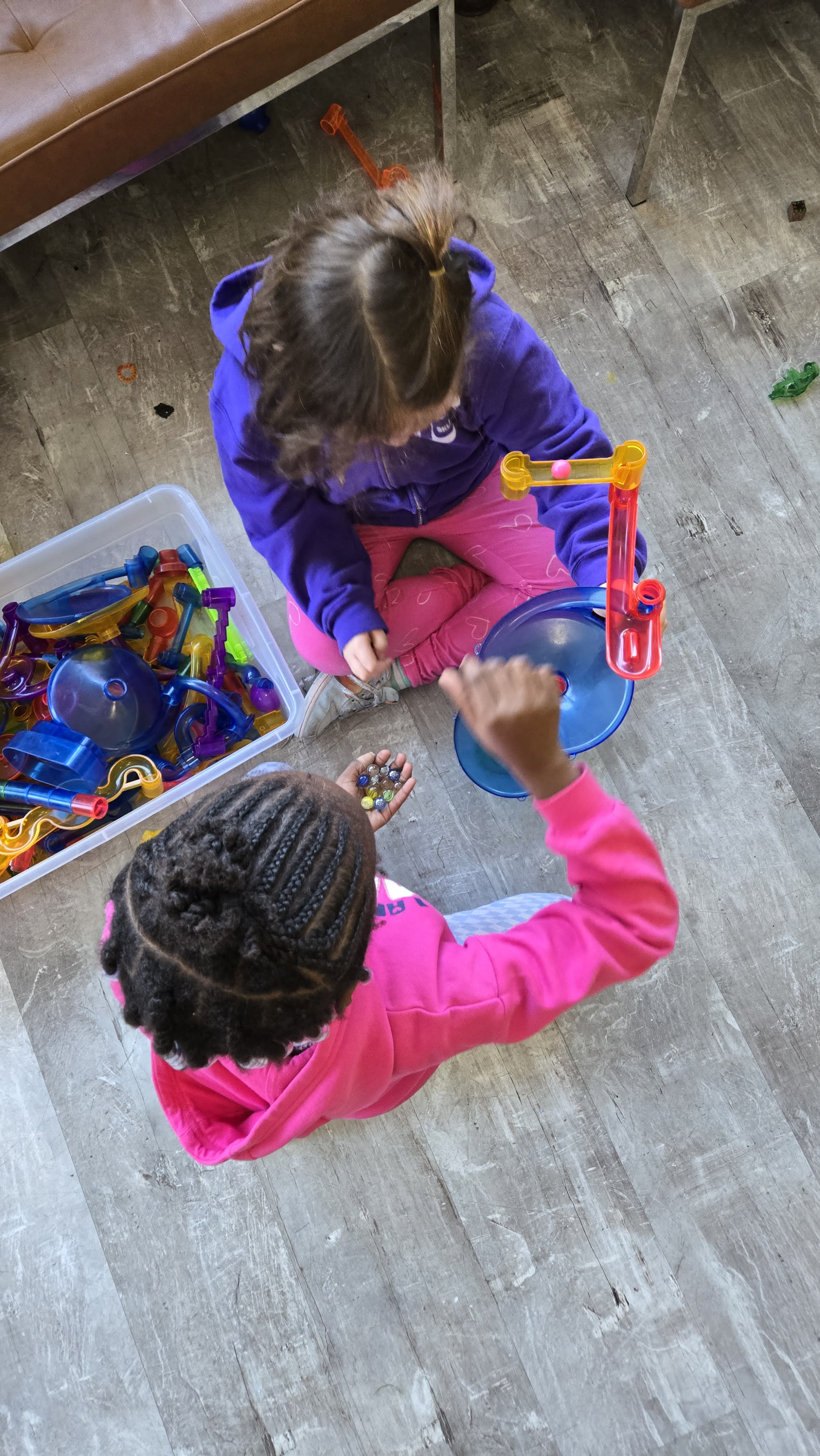 Two children sitting on the floor playing with a marble run toy and a container of colorful parts.