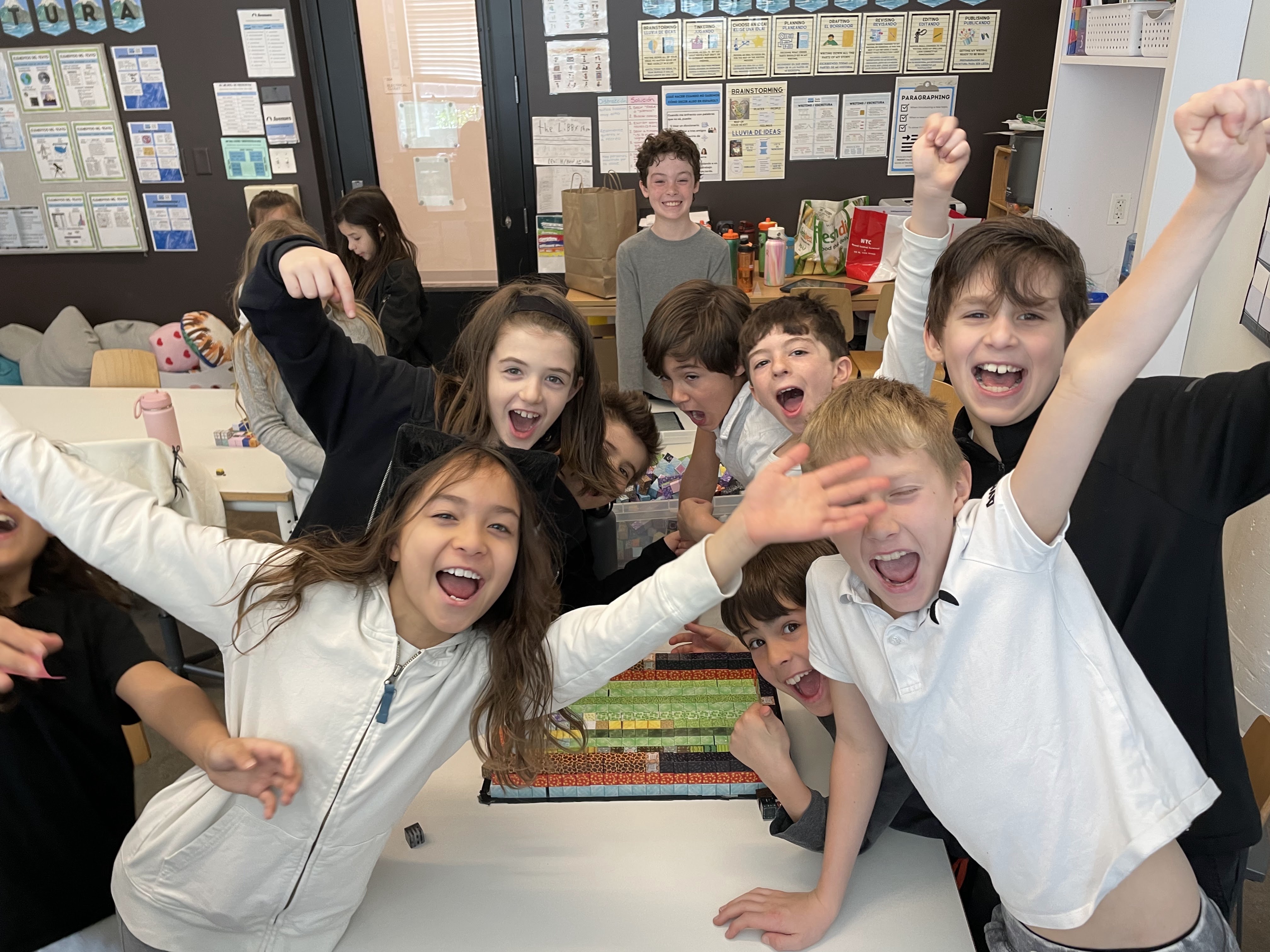 Group of children smiling and cheering around a colorful block project on a classroom table.