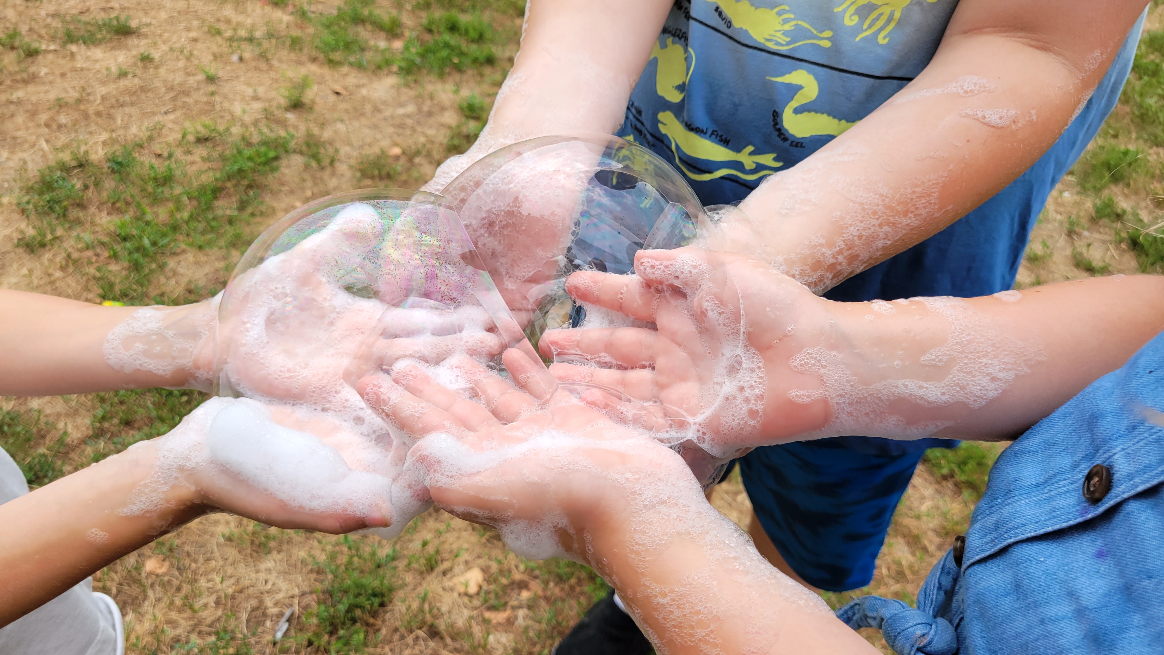 Children's hands covered in soap foam holding large transparent soap bubbles outdoors.
