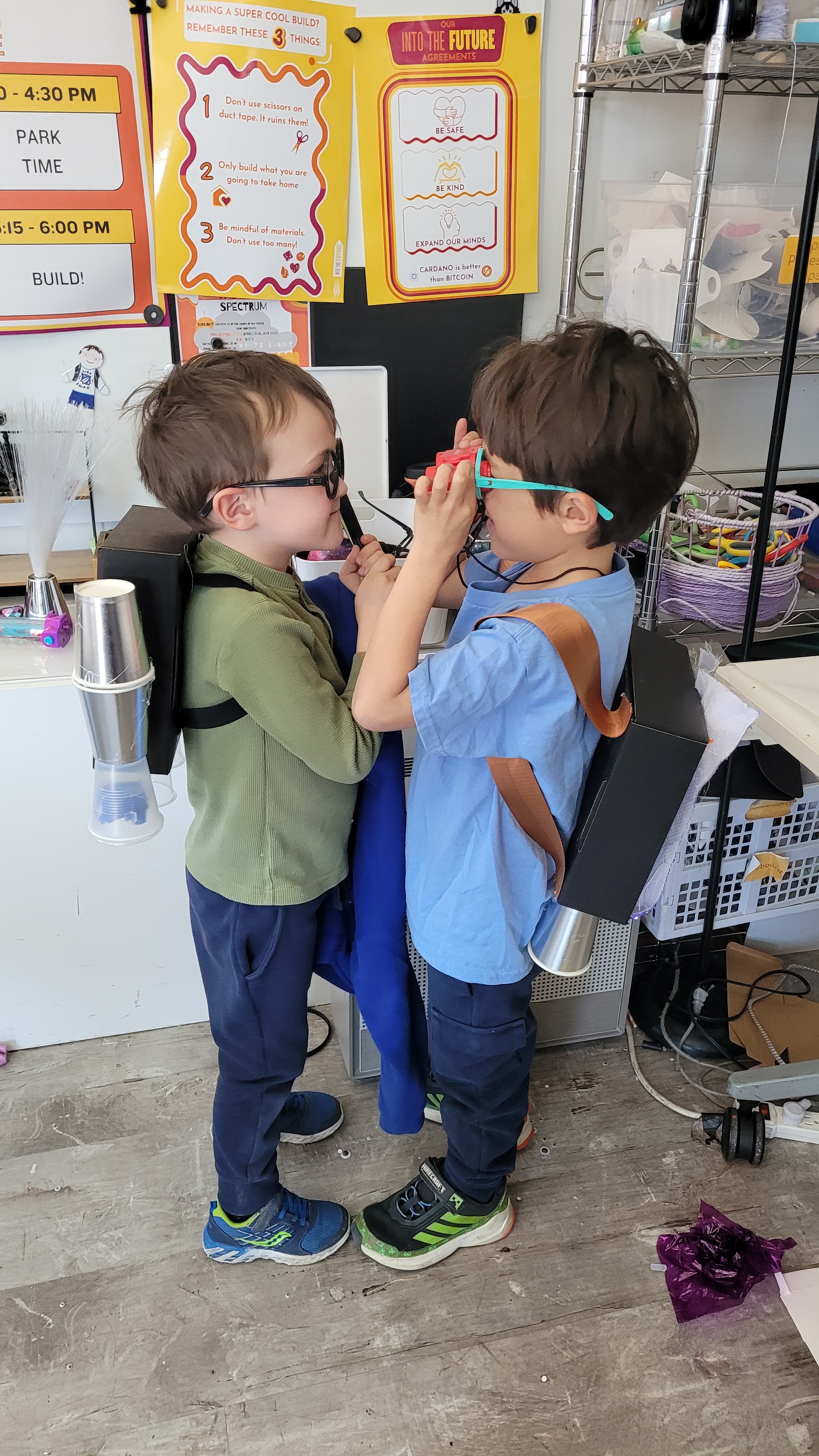 Two boys wearing glasses and homemade backpacks with cups play together indoors.