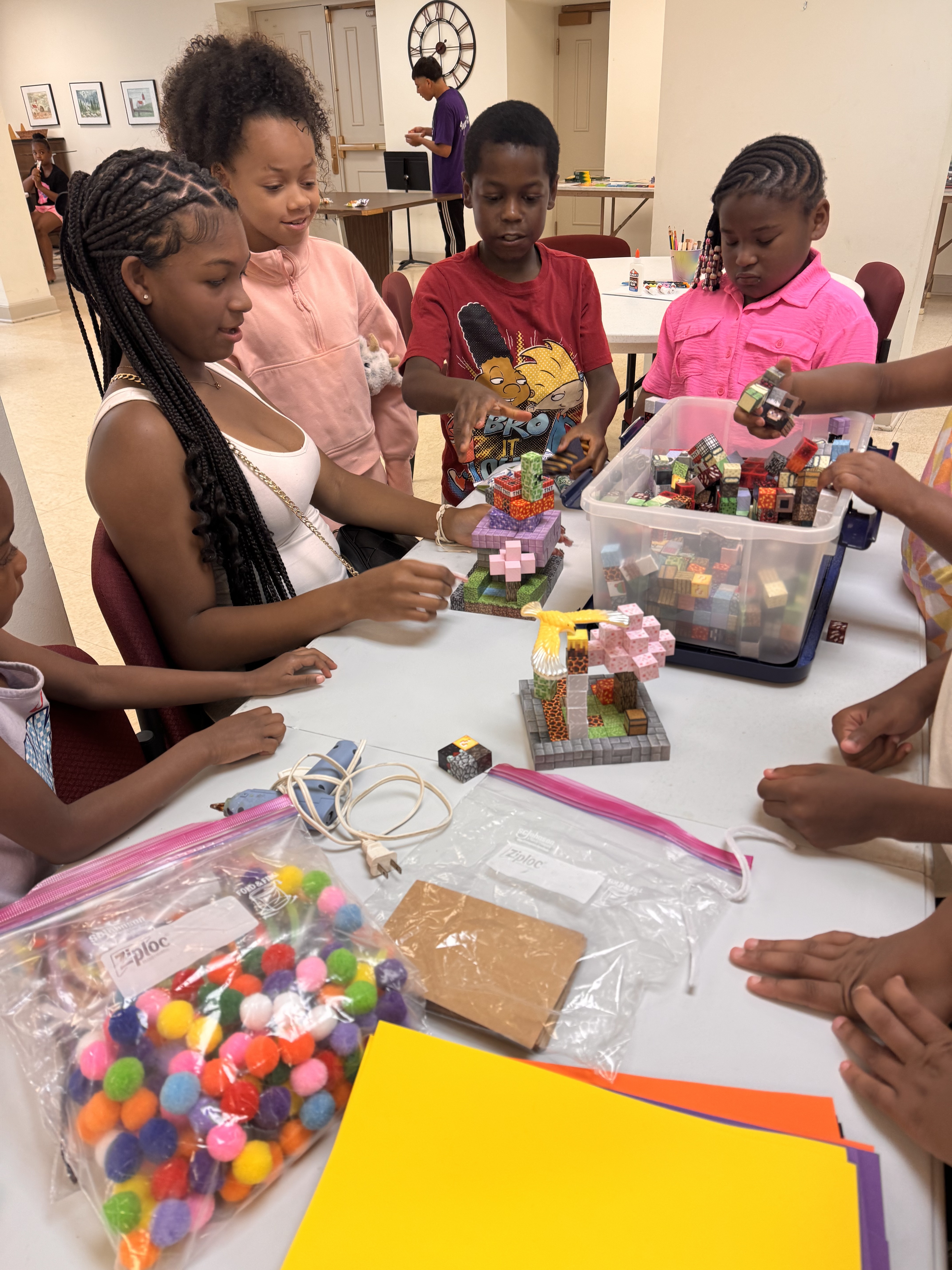 Children and a teen build block structures at a table with arts and crafts supplies around them.