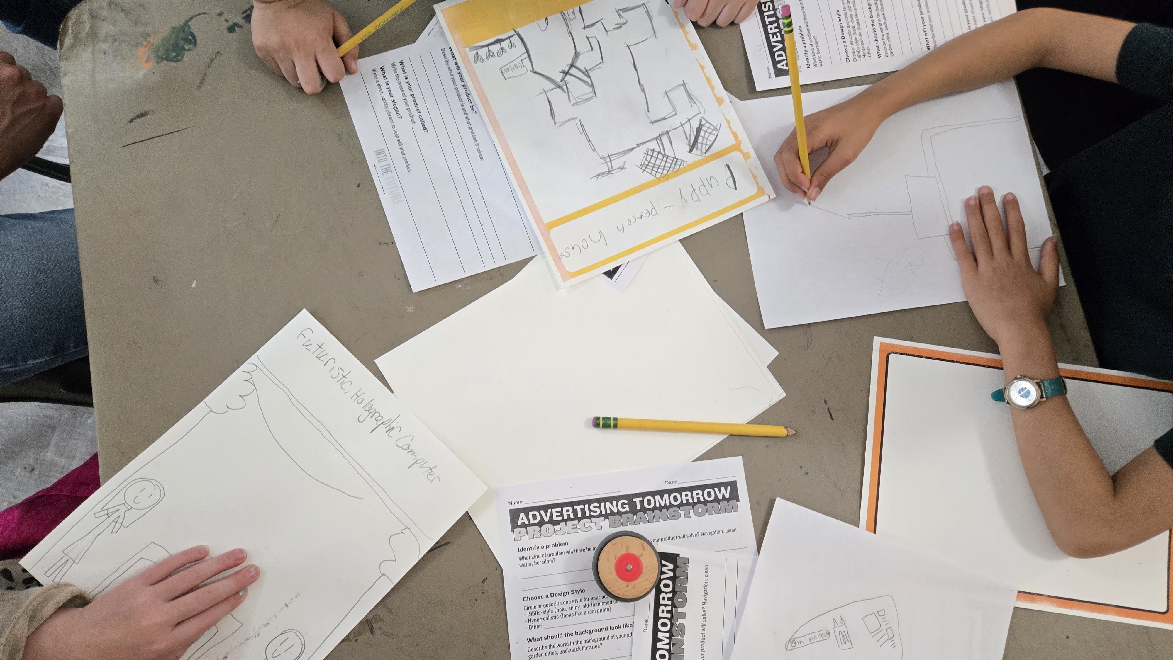 Children sketching and writing on papers around a table labeled Advertising Tomorrow project brainstorm.