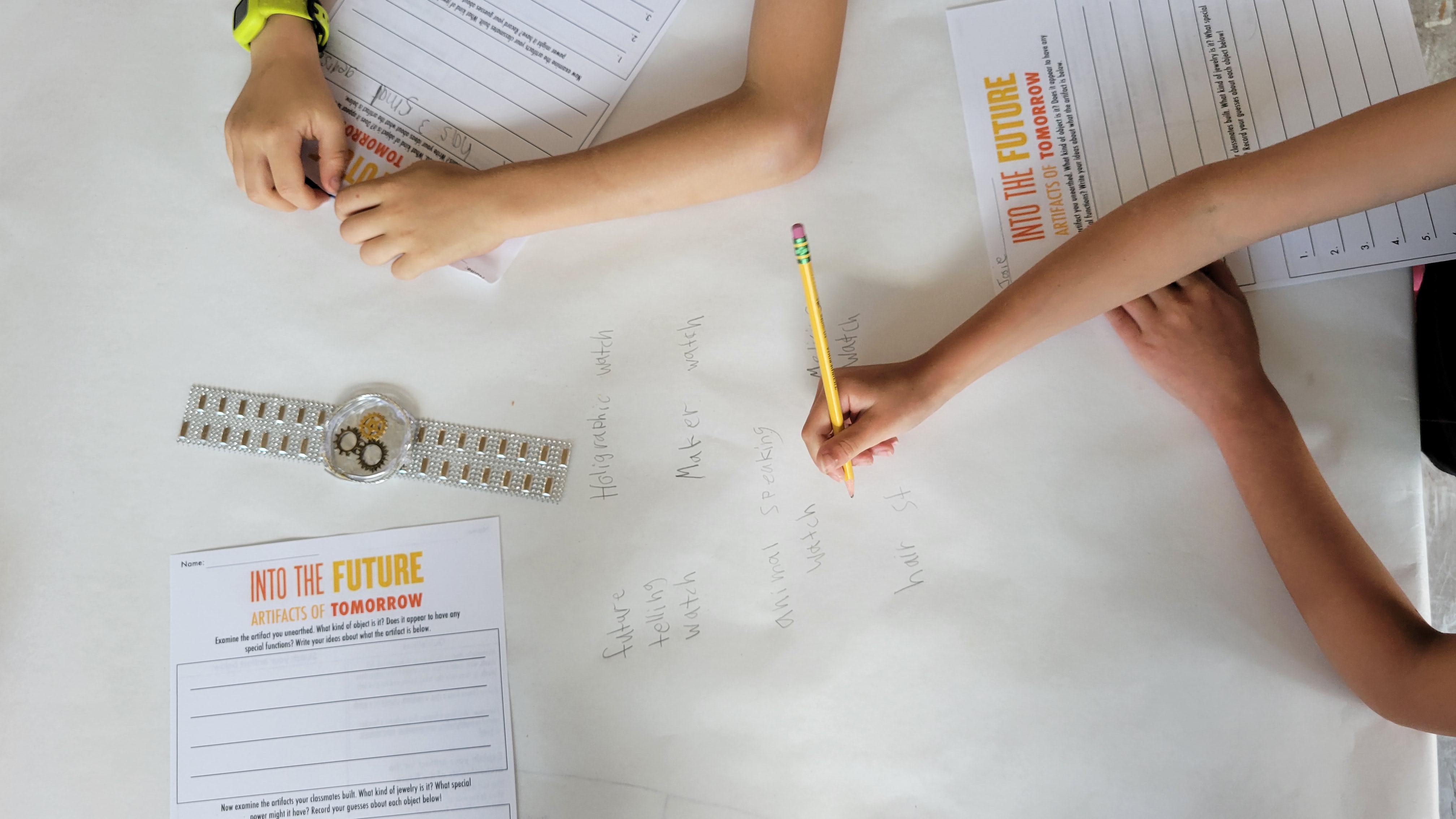Two children writing ideas and drawing futuristic watches on large paper with worksheets nearby.