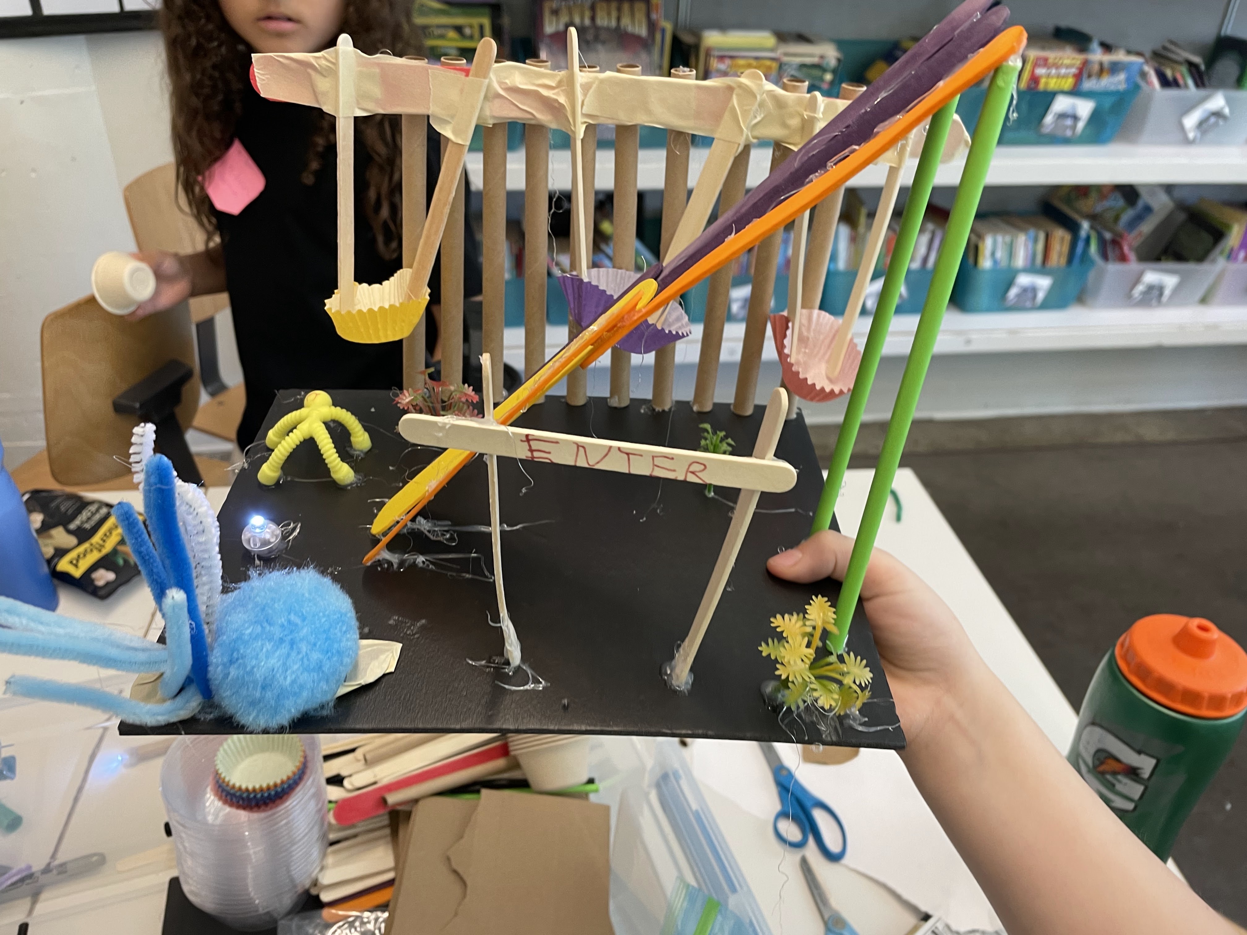 Child holding a craft model of a colorful miniature playground with swings and an 'ENTER' sign.