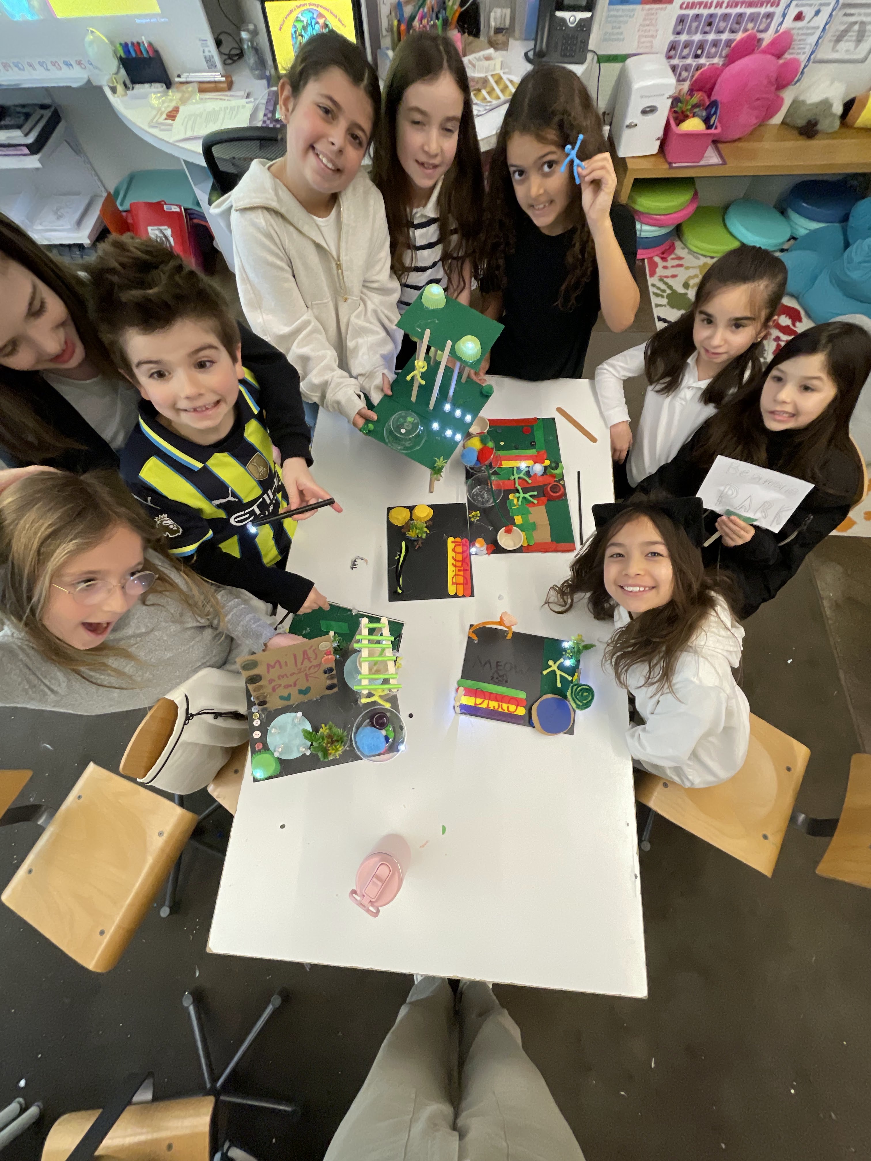 Group of children smiling around a table showing handmade craft projects in a classroom setting.