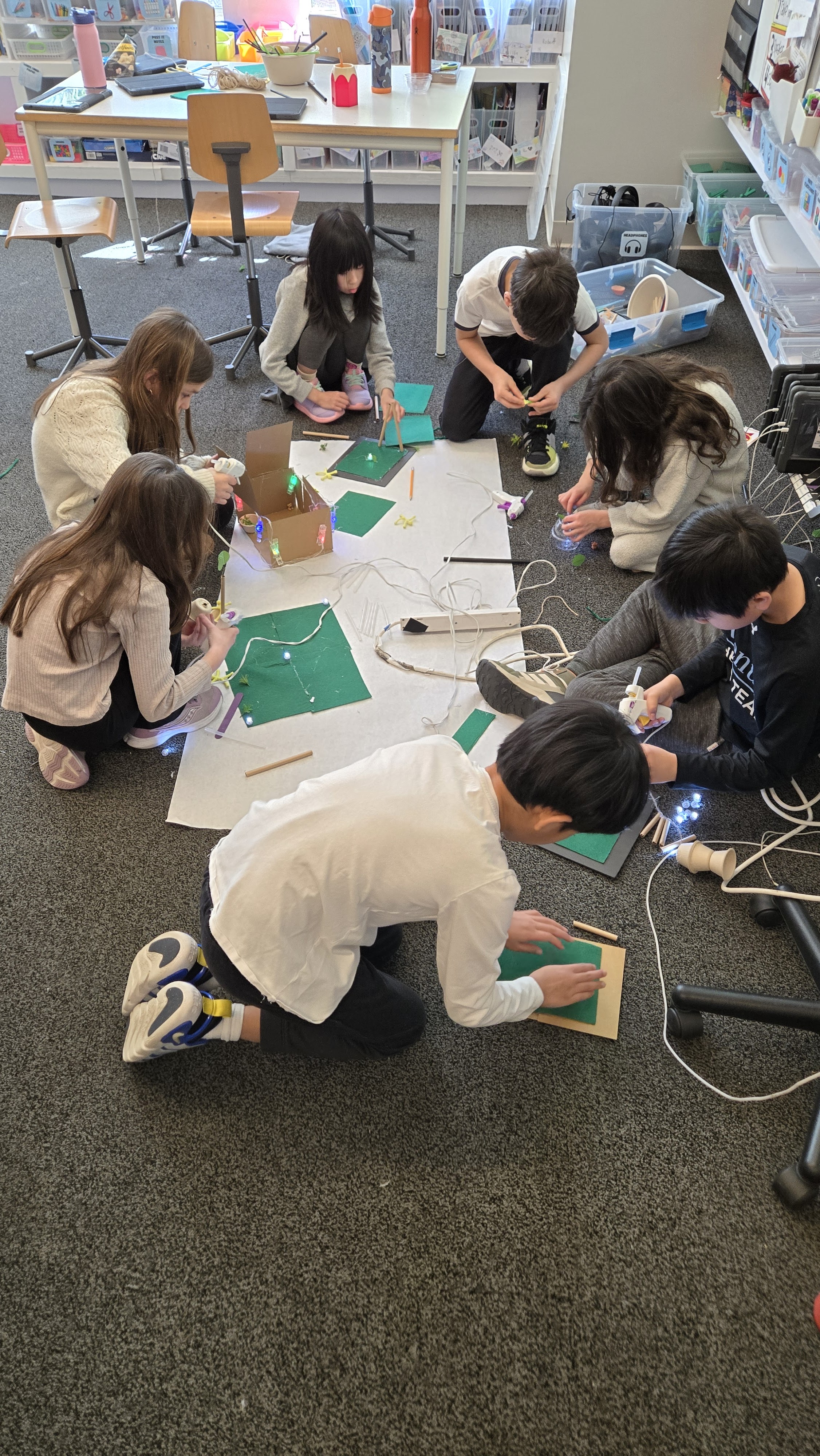 Seven children sitting and kneeling on a classroom floor working on a craft project with green felt and lights.
