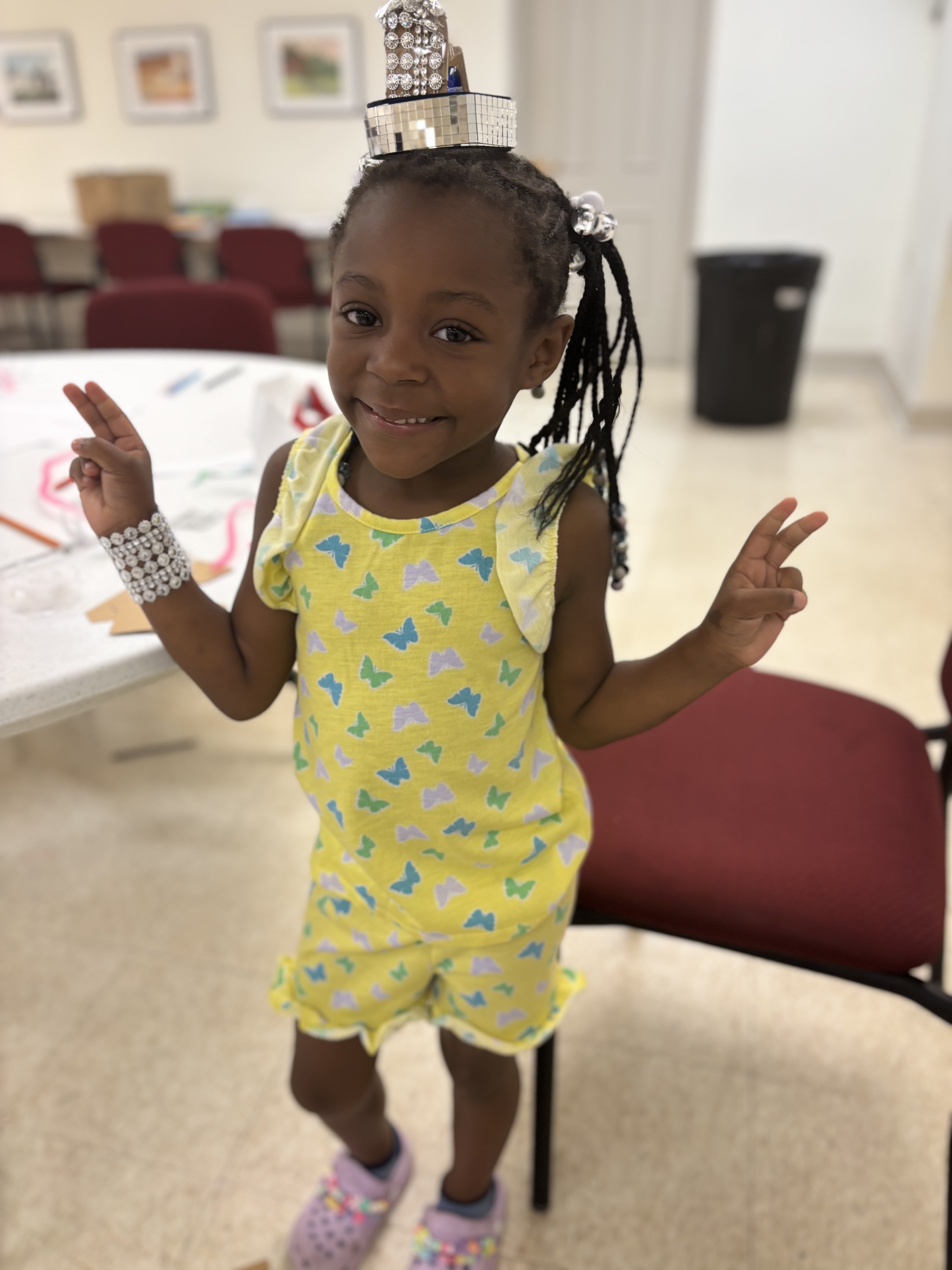 Smiling girl in yellow butterfly outfit and beaded accessories flashes peace signs in a room with chairs and table.
