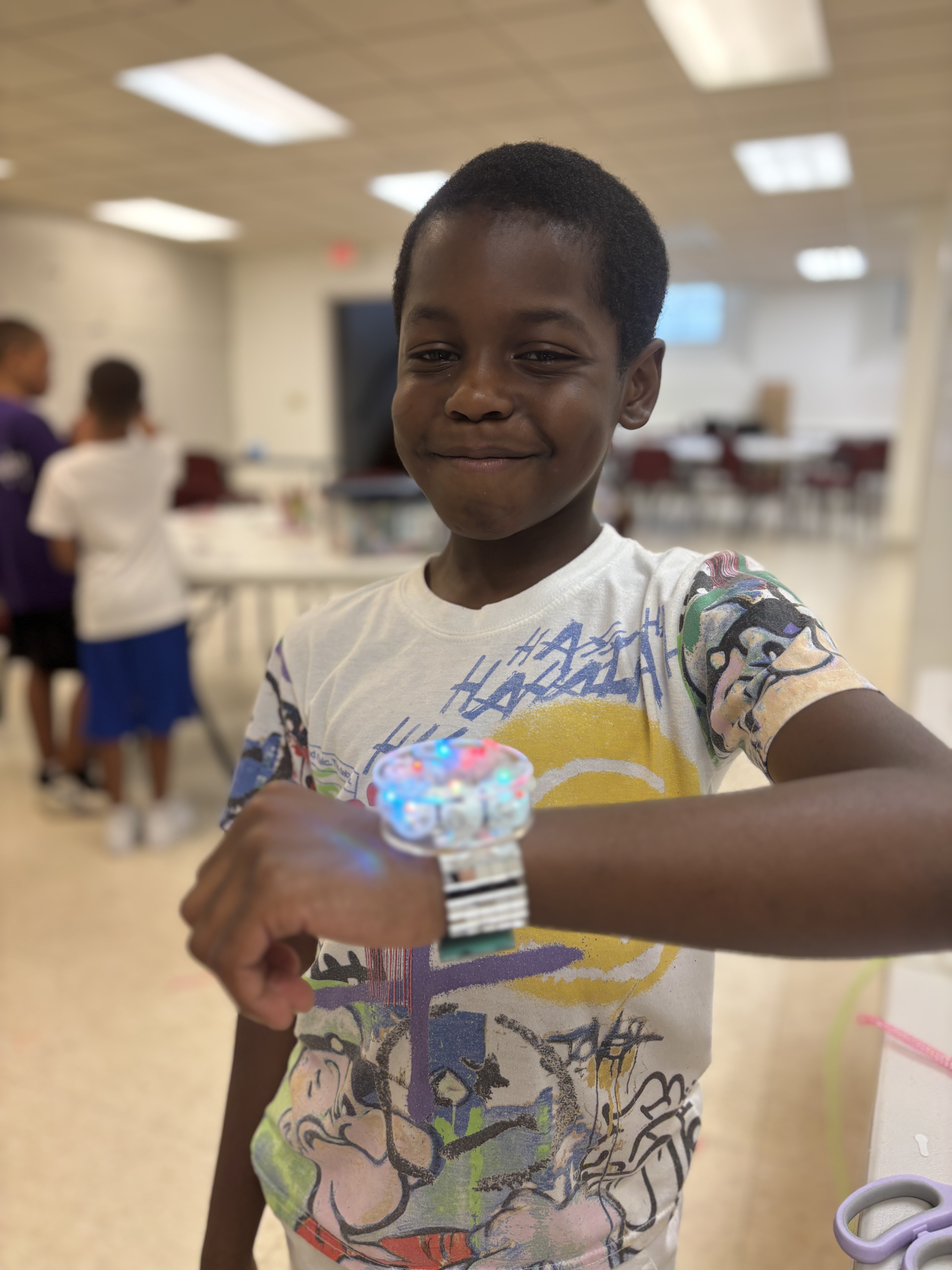 Smiling boy wearing a colorful t-shirt showing a bright, colorful bracelet on his wrist indoors.