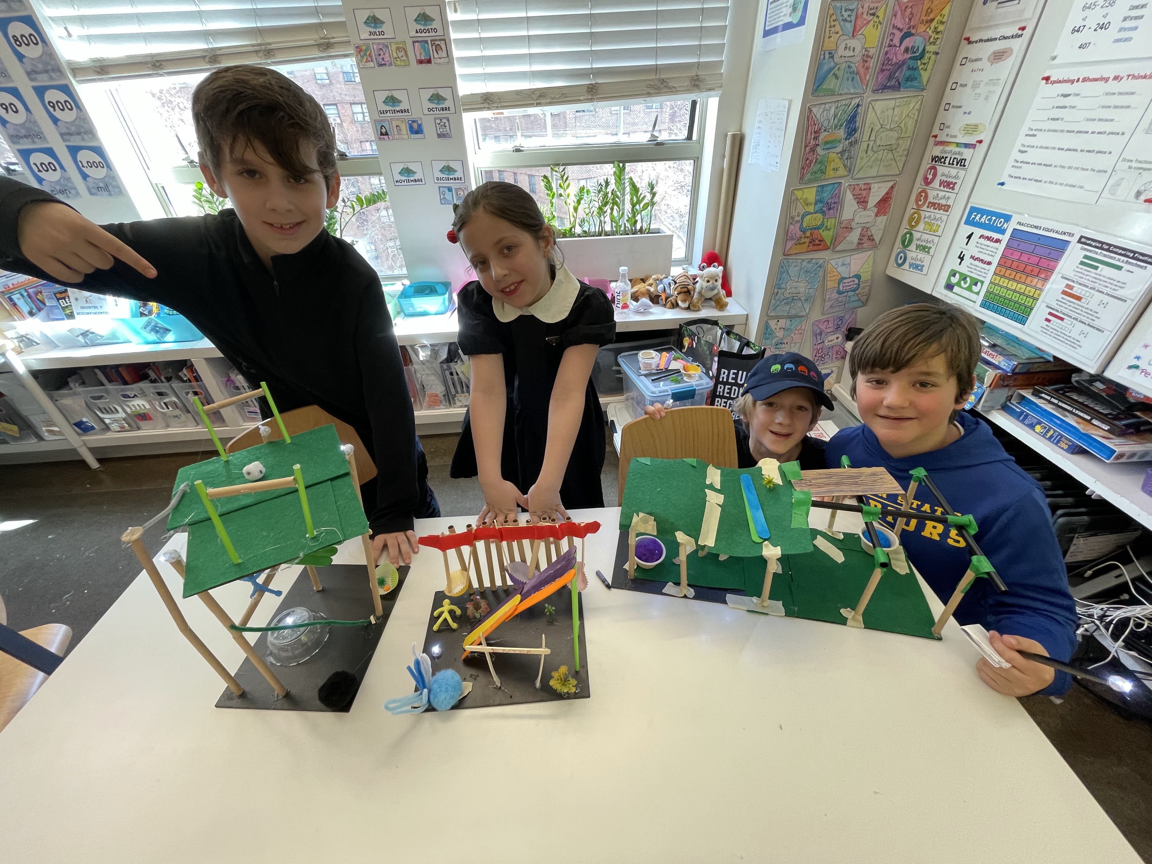 Four children in a classroom showing handmade craft projects on a table, smiling at the camera.