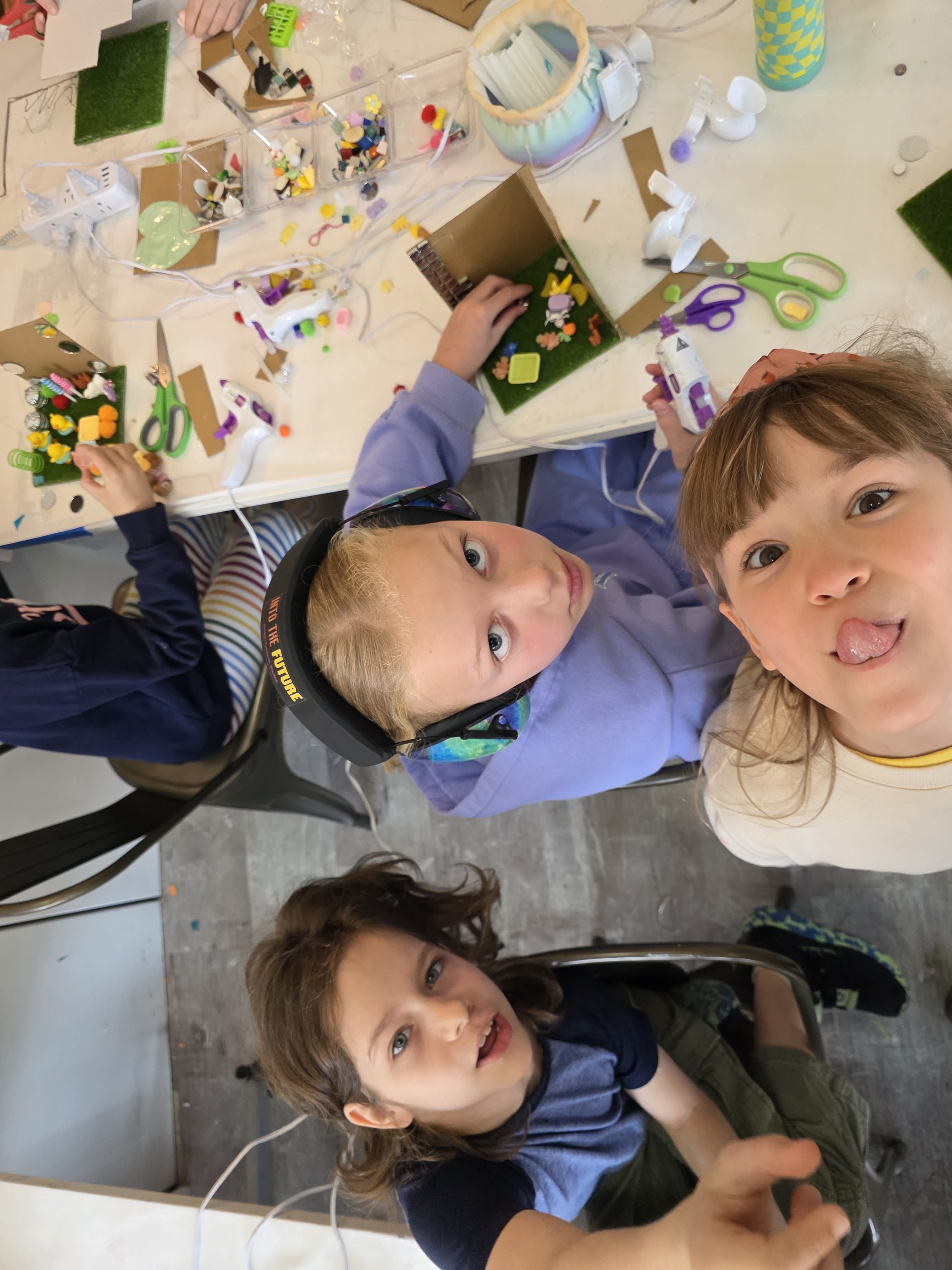 Three children making crafts with glue guns and colorful materials at a cluttered table.