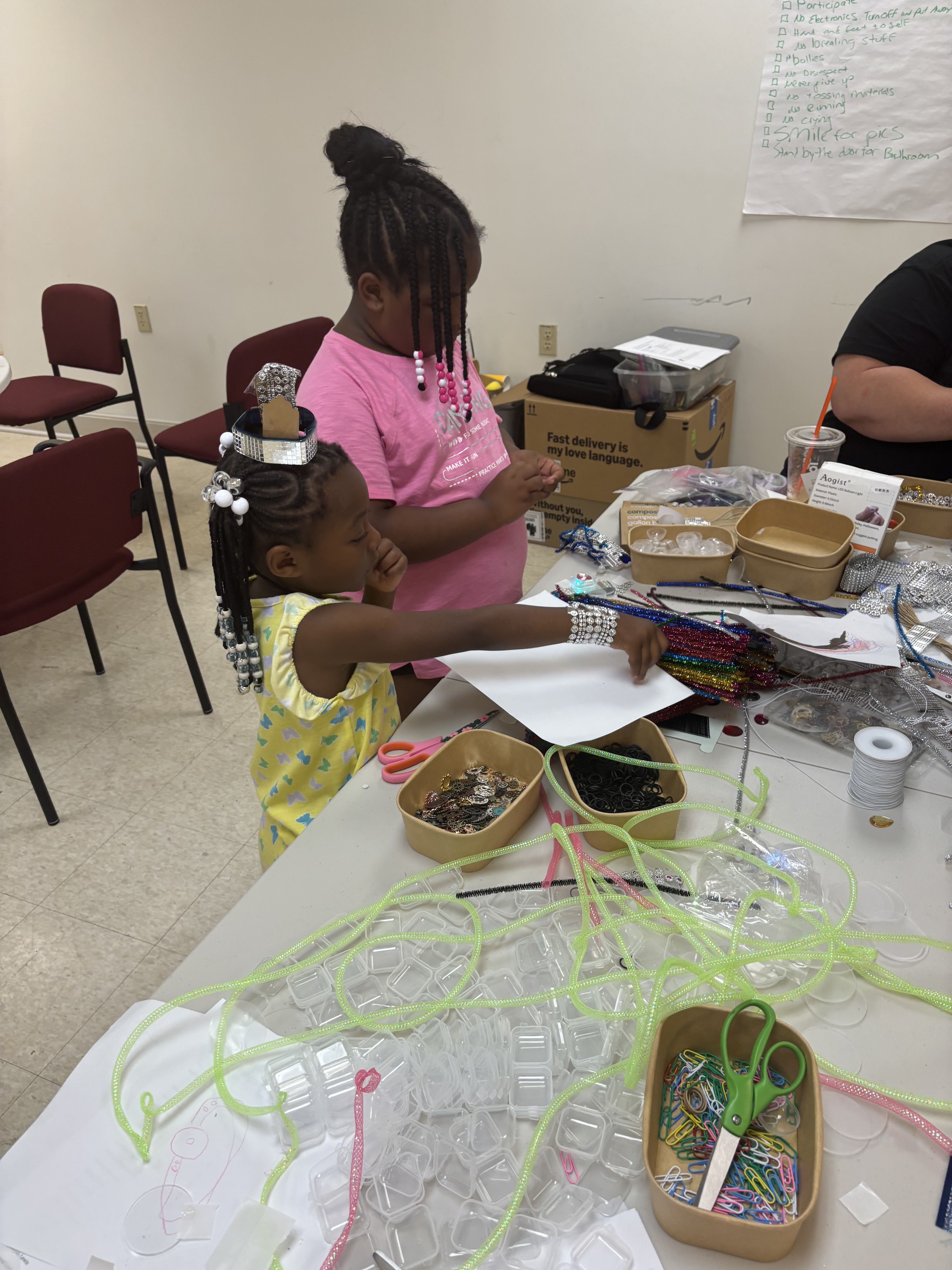 Two girls crafting at a table with beads, scissors, paper, and colorful craft supplies.