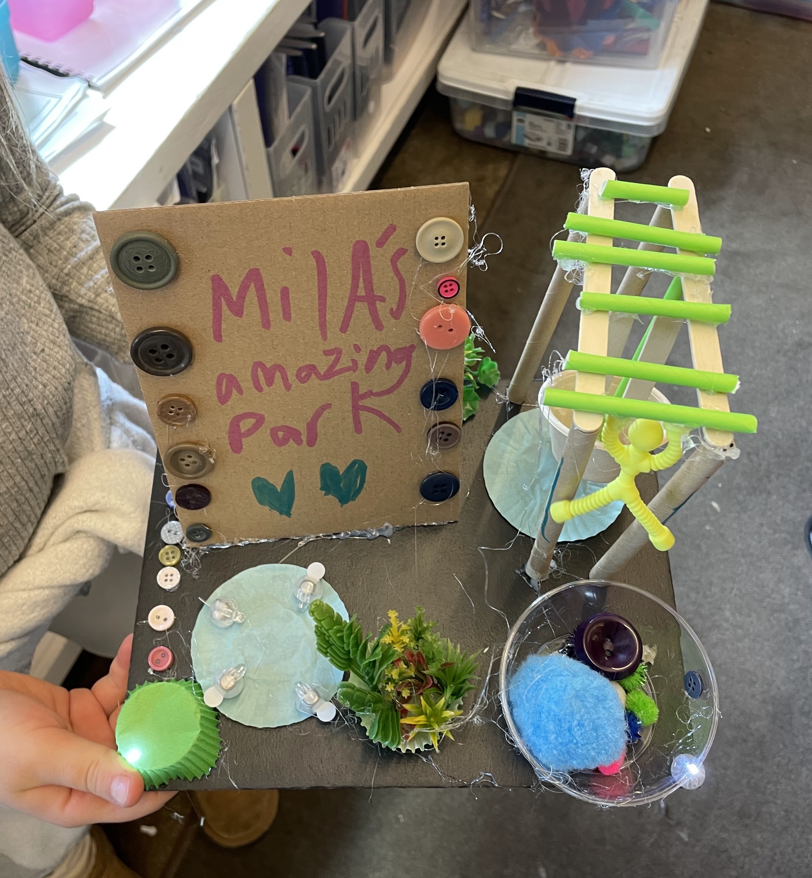 Handmade miniature park model with ladder, plants, lights, buttons, and sign saying Milas amazing park.