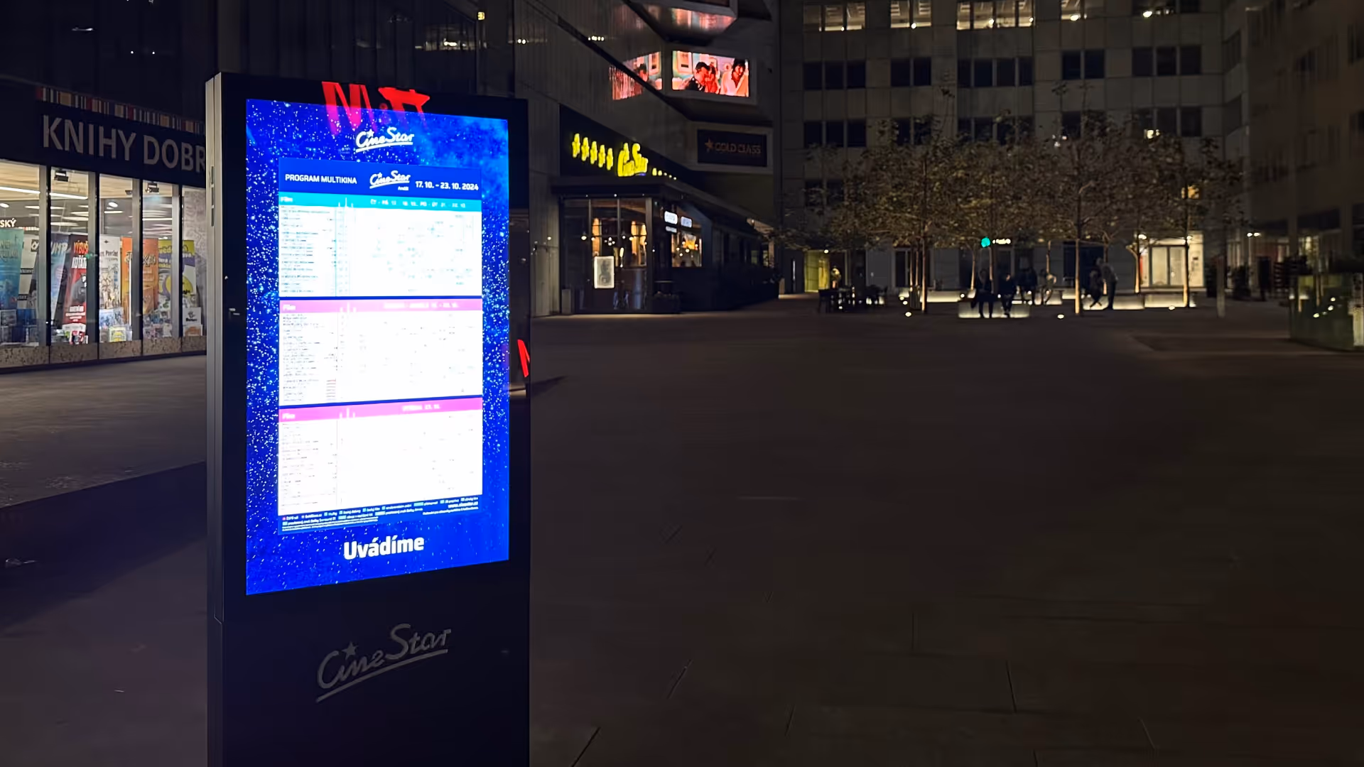 Illuminated CineStar movie schedule display board at night on an open plaza with buildings and people in the background.