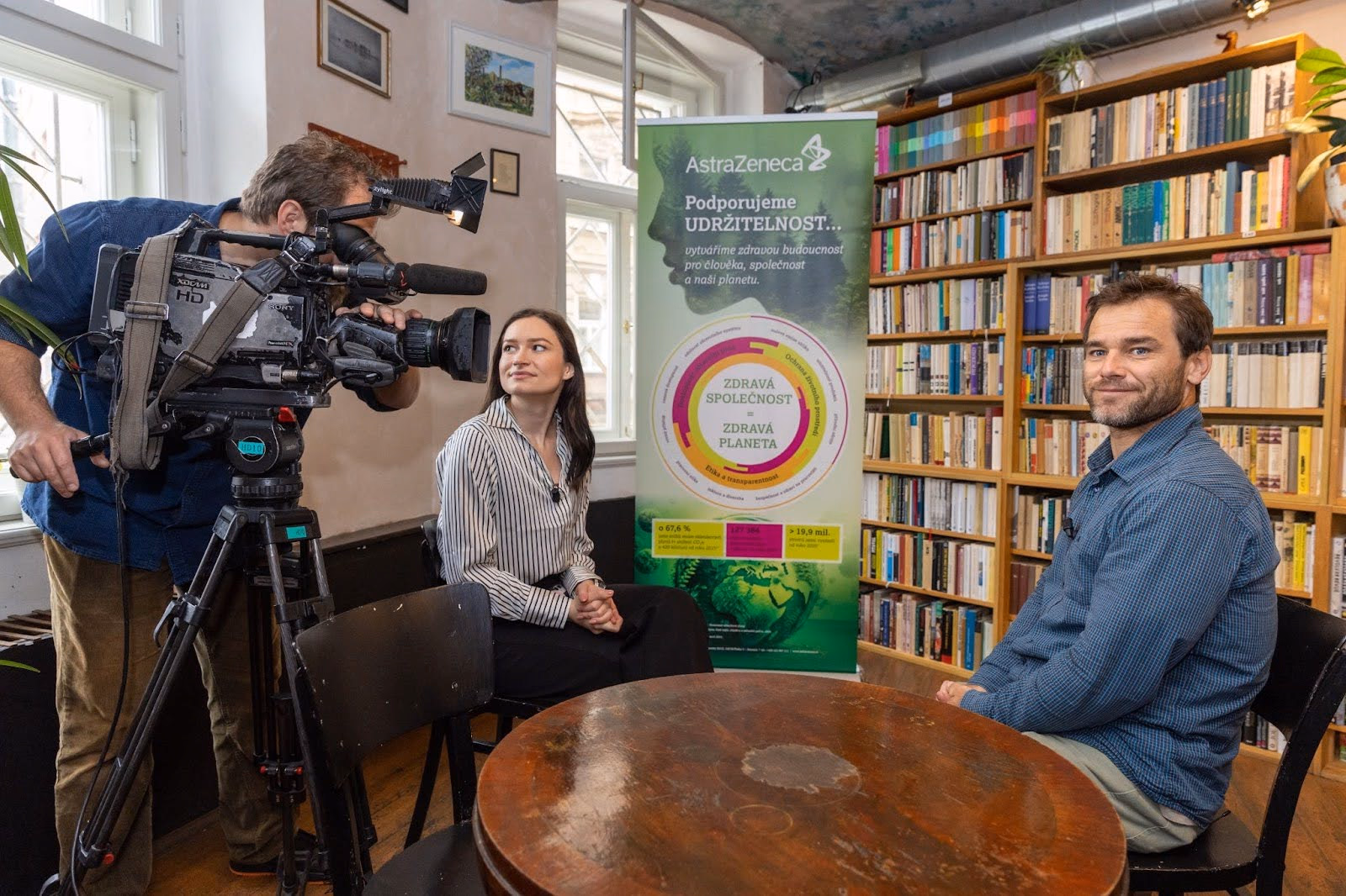 A man with a video camera films a woman sitting at a table with a man in a room filled with bookshelves and an AstraZeneca sustainability poster in the background.