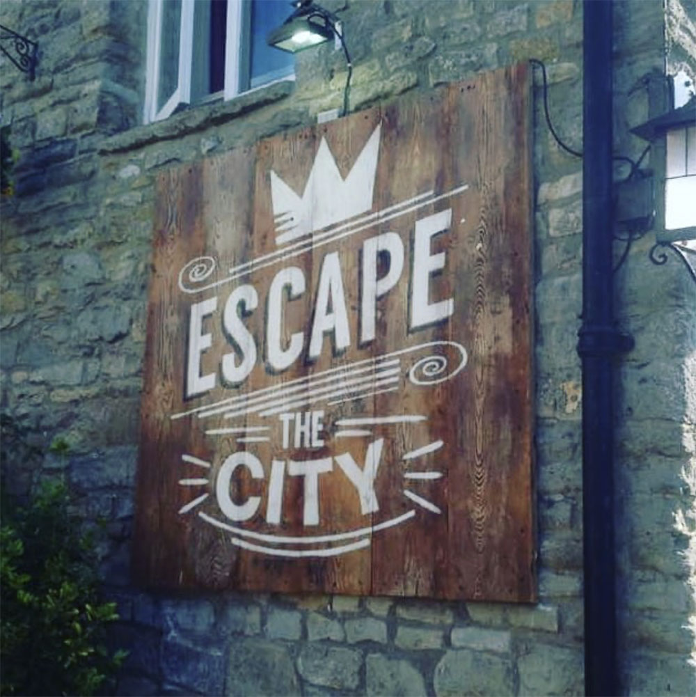 Wooden sign with white text 'Escape the City' mounted on a stone wall under an outdoor light.