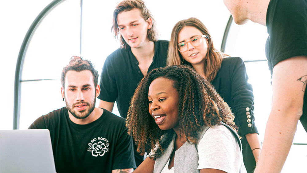 Group of five diverse young adults collaborating around a laptop in a bright room with arched windows.