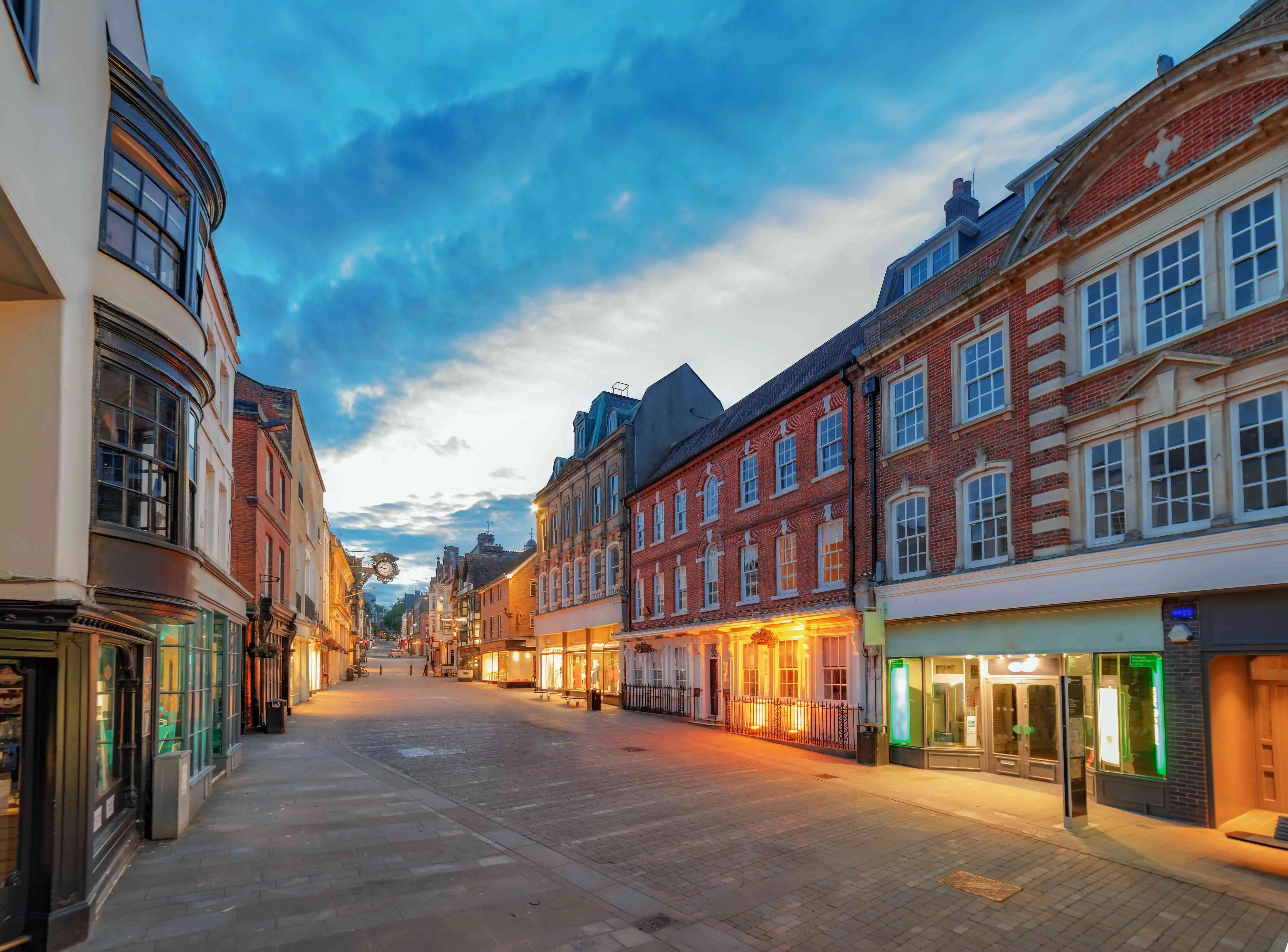 Empty pedestrian street lined with historic brick and stone buildings at dusk with glowing shop windows under a cloudy blue sky.