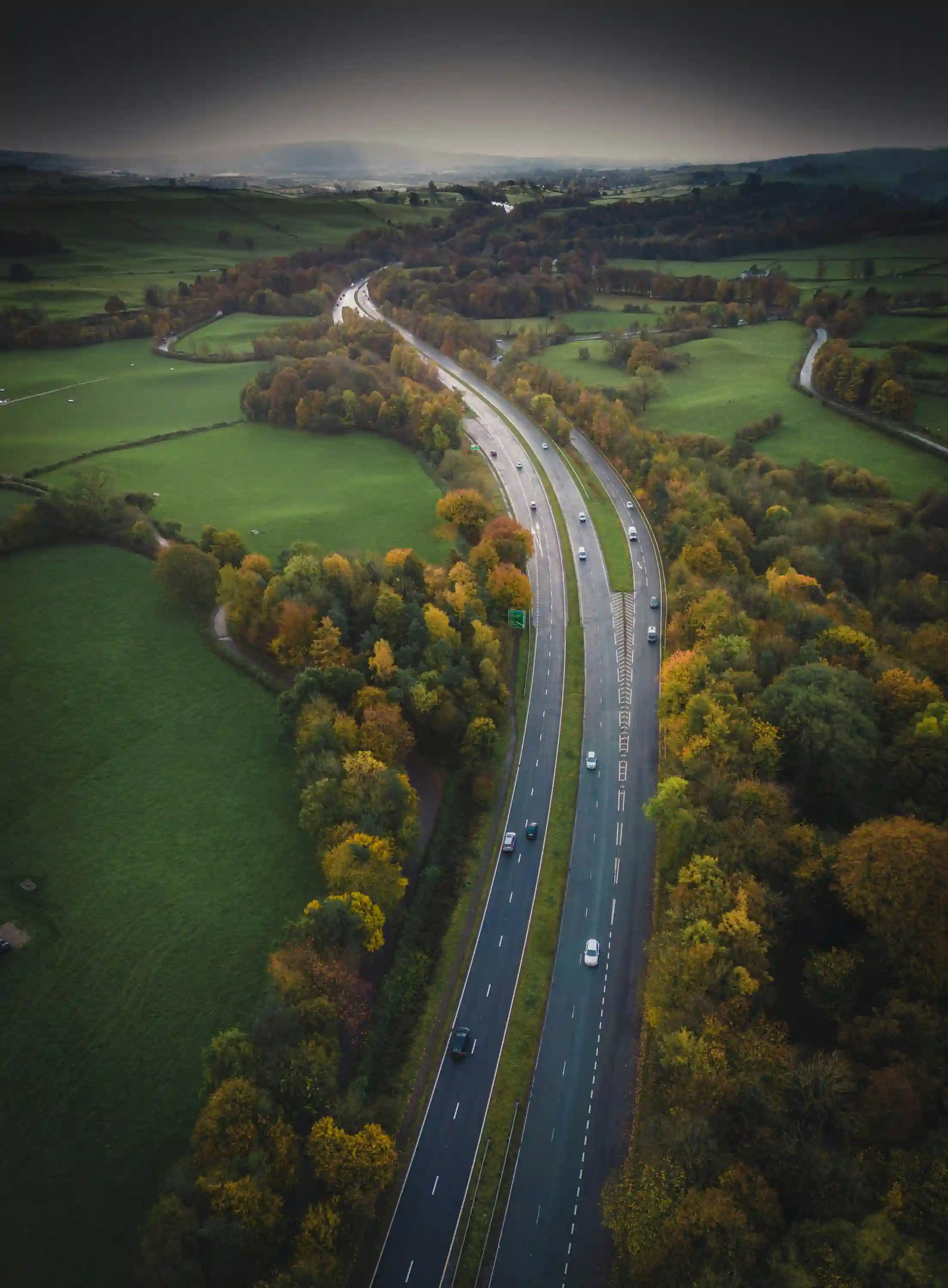 Aerial view of a winding highway surrounded by green fields and trees with autumn foliage.