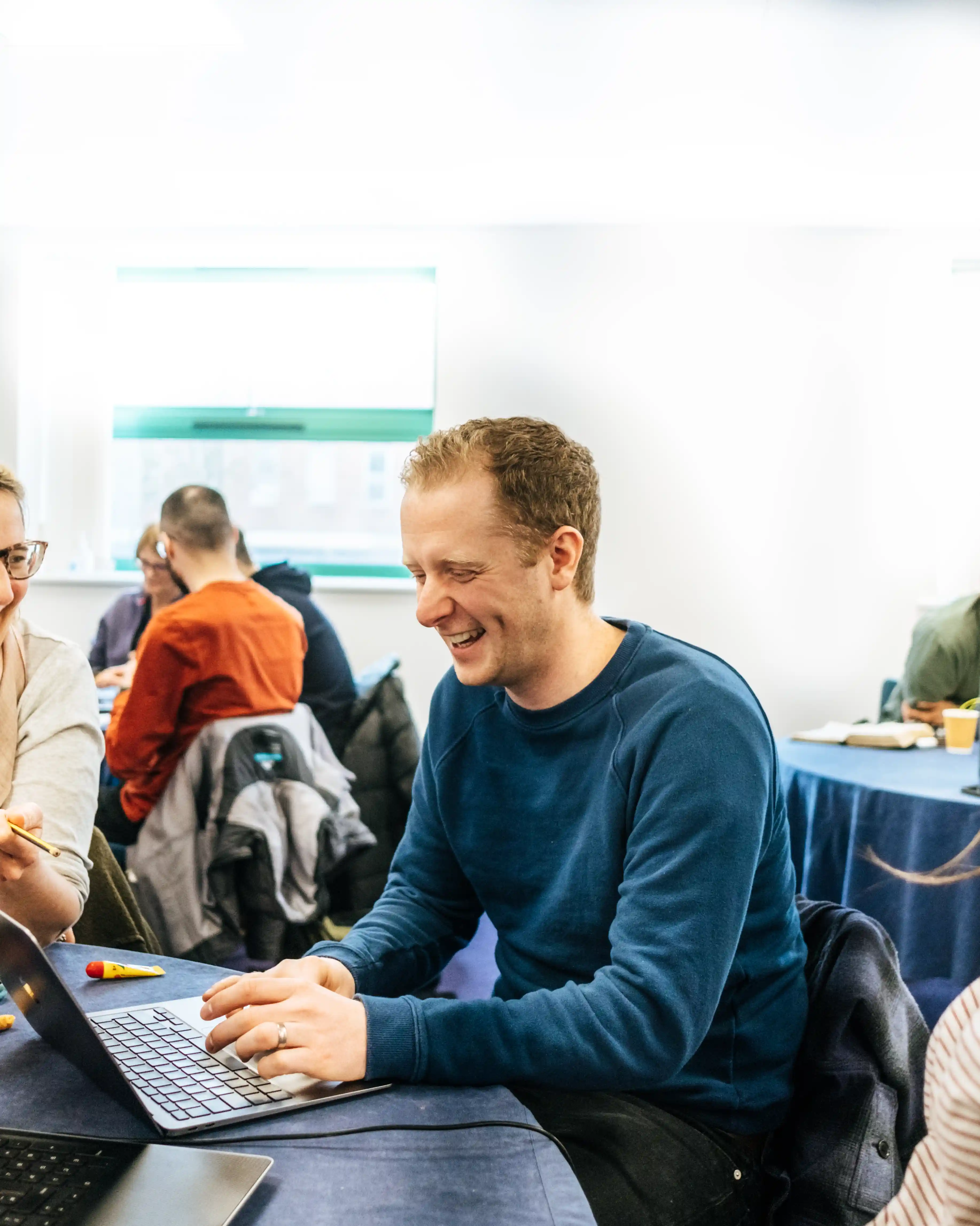 Man in blue sweater laughing and typing on a laptop at a table in a bright room with others working in the background.