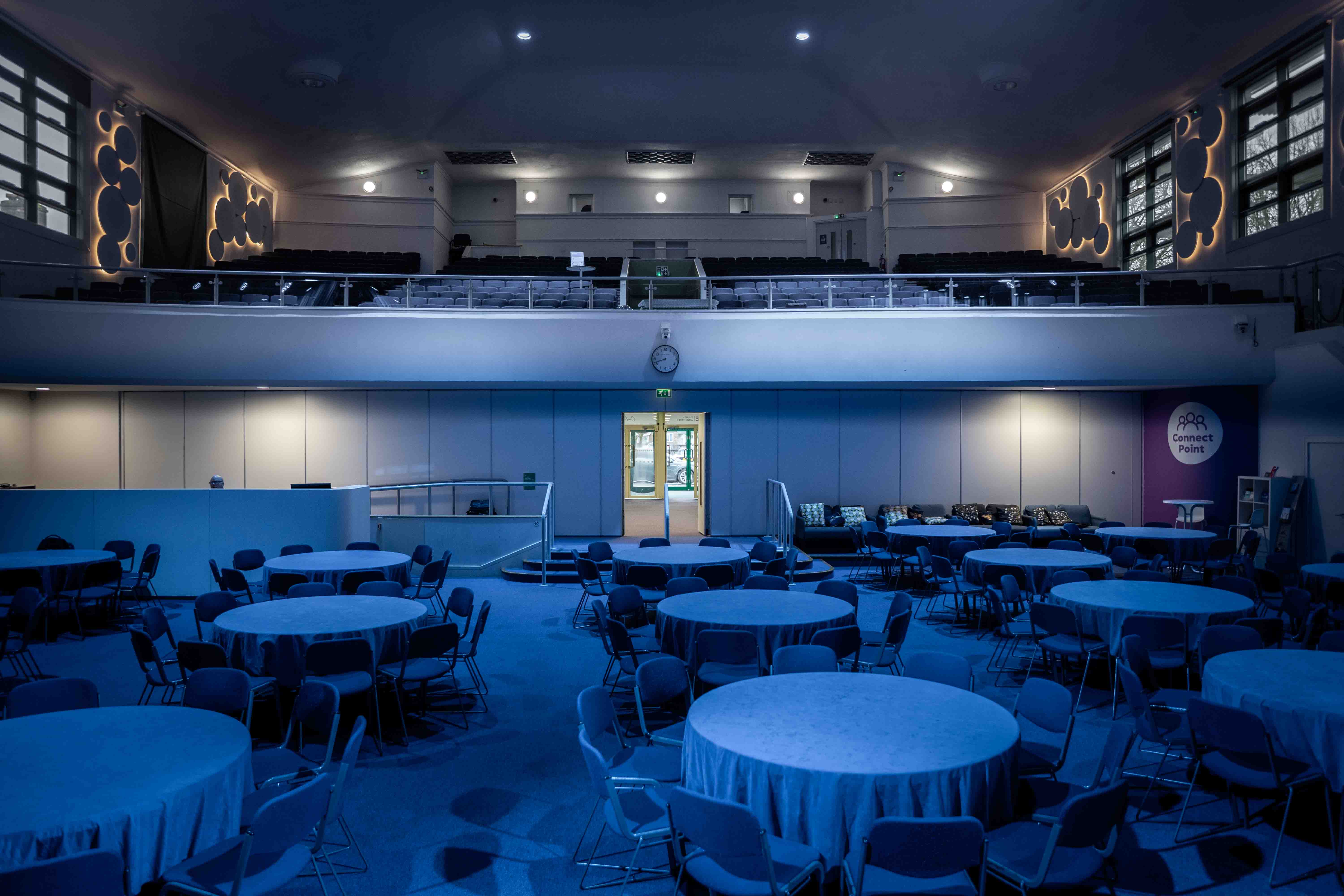 Interior view of a large event space with round tables covered in cloth, surrounded by chairs, blue carpet, mezzanine seating, and a door at the back wall.