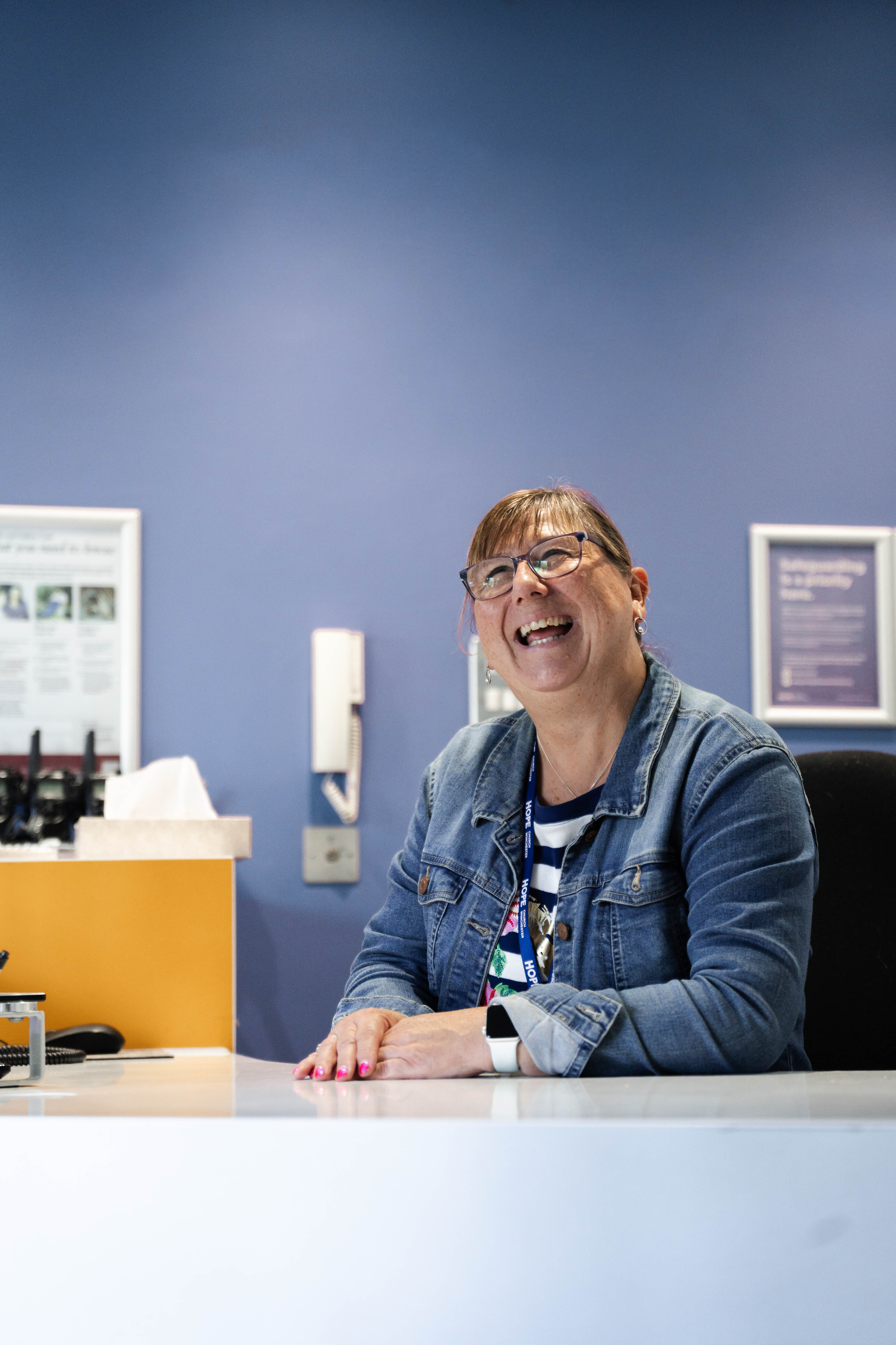 Smiling woman wearing glasses and a denim jacket sitting at a white reception desk against a blue wall.