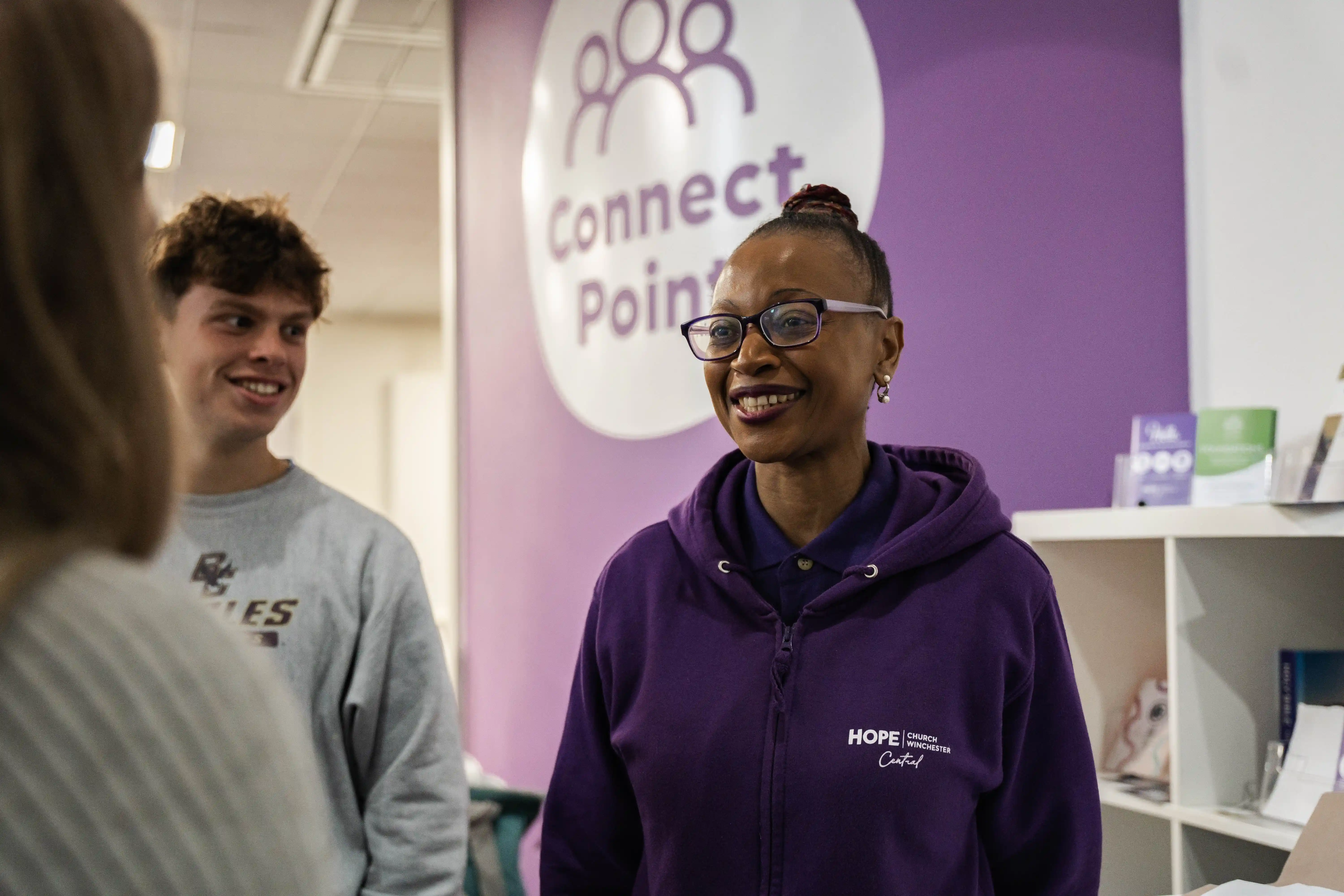 Smiling woman in a purple hoodie with 'HOPE Church Winchester Central' talks to two people at Connect Point desk.