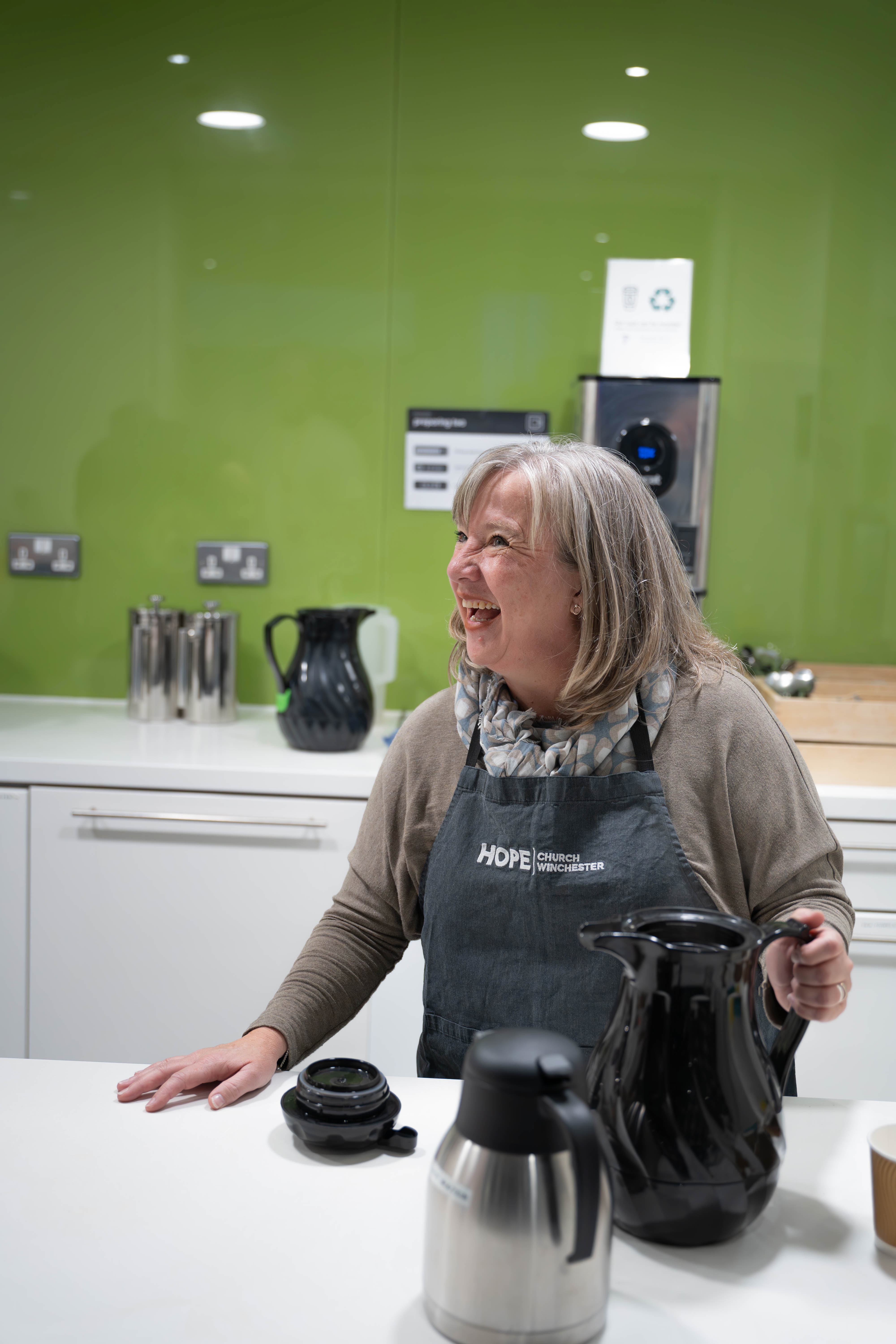 Smiling woman in a Hope Church Winchester apron standing behind a kitchen counter with coffee pots and a green wall background.