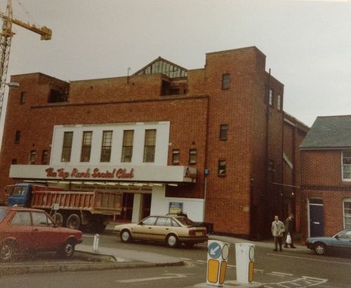 Vintage street scene with a large brick building labeled 'The Top Rank Social Club,' parked cars, a construction truck, and two people walking on the sidewalk.