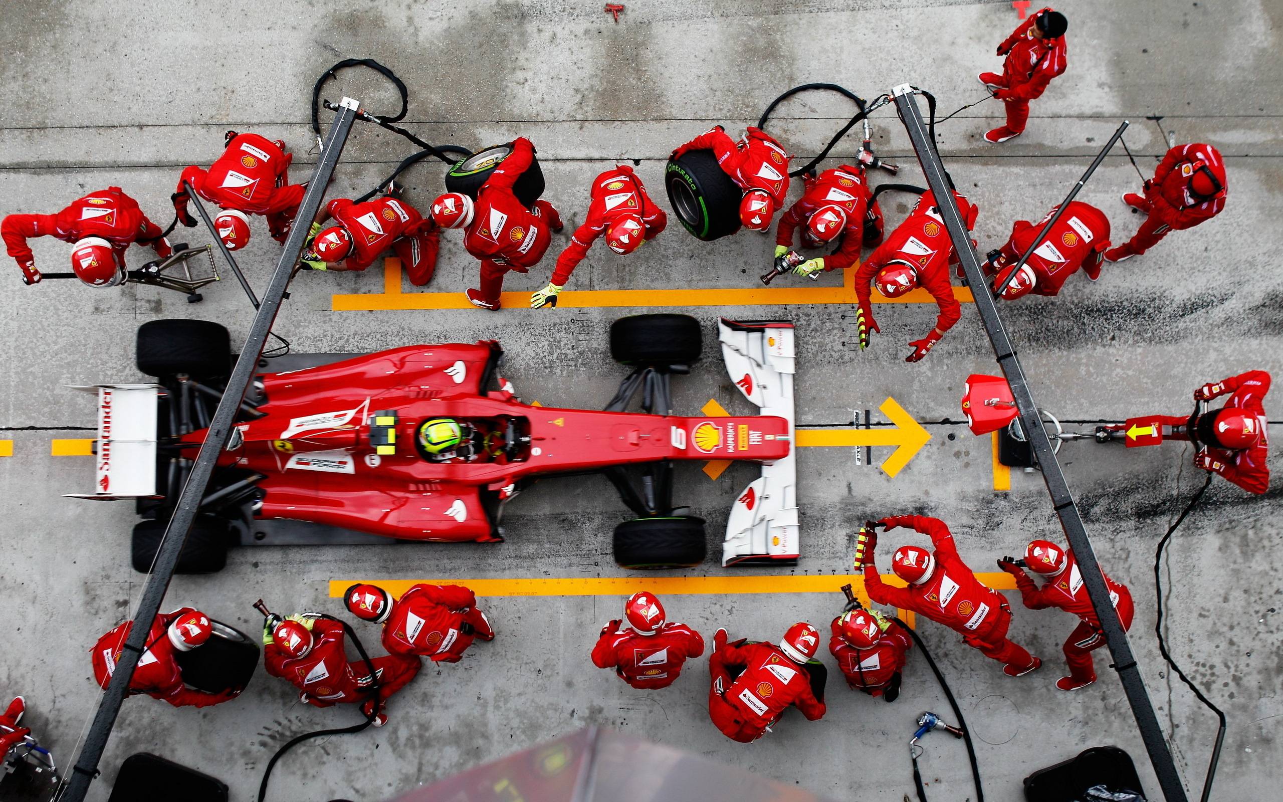 Aerial view of a Formula 1 pit stop showing a red race car surrounded by a coordinated pit crew in red uniforms performing maintenance and tire changes.