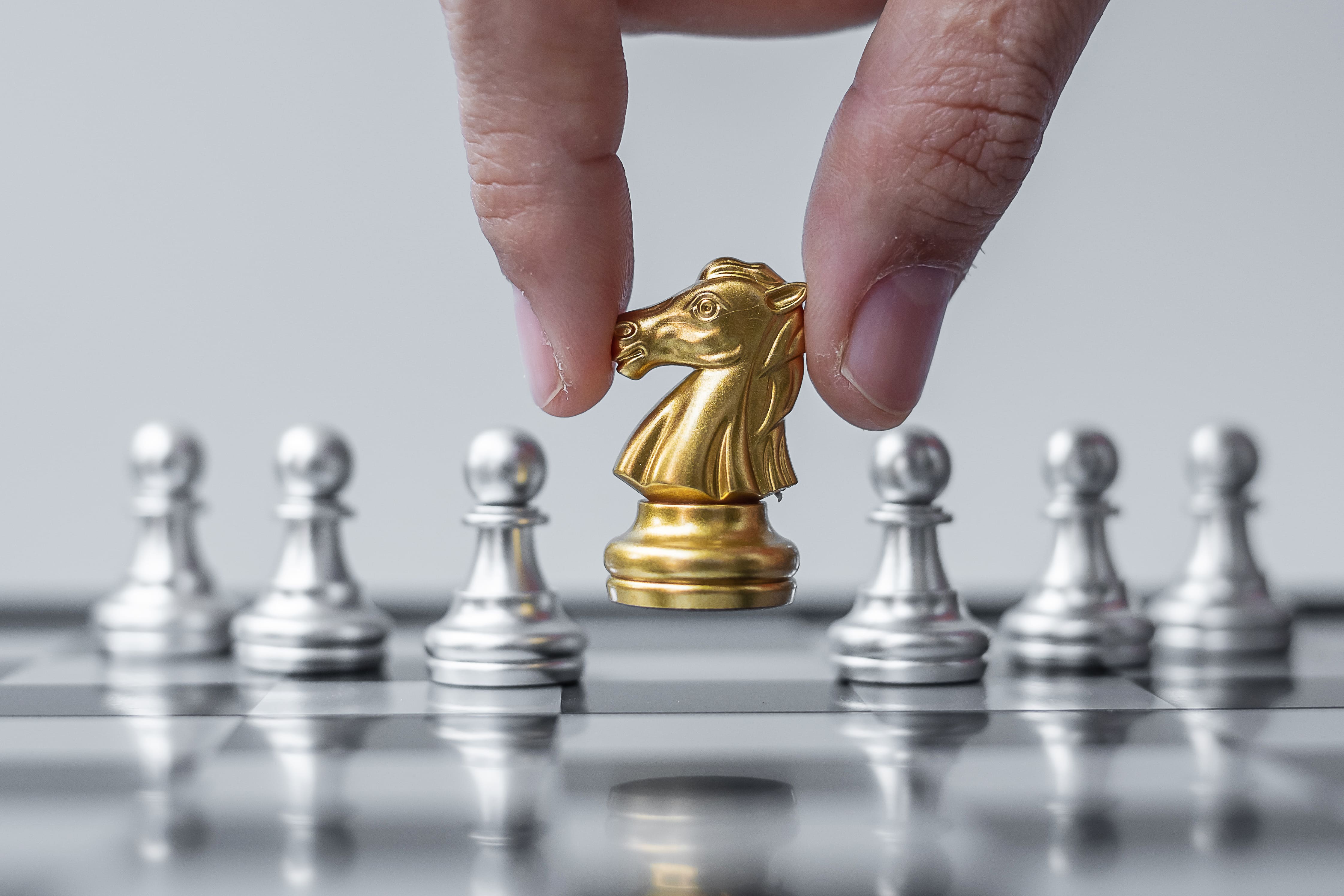 Close-up of a hand placing a golden knight chess piece among silver pawns on a chessboard, symbolizing strategy, leadership, and innovation.