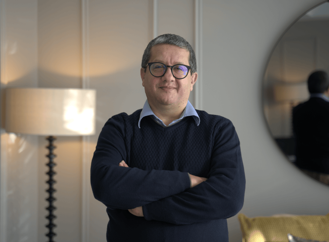 Man wearing glasses, a navy sweater, and a light blue shirt standing with arms crossed in a warmly lit room with a lamp and round mirror in the background.