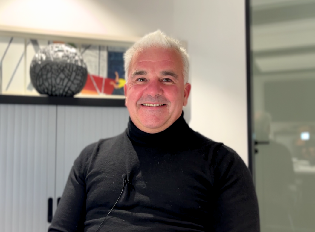 Smiling man with short white hair wearing a black turtleneck sweater, seated indoors with modern decor and artwork in the background.