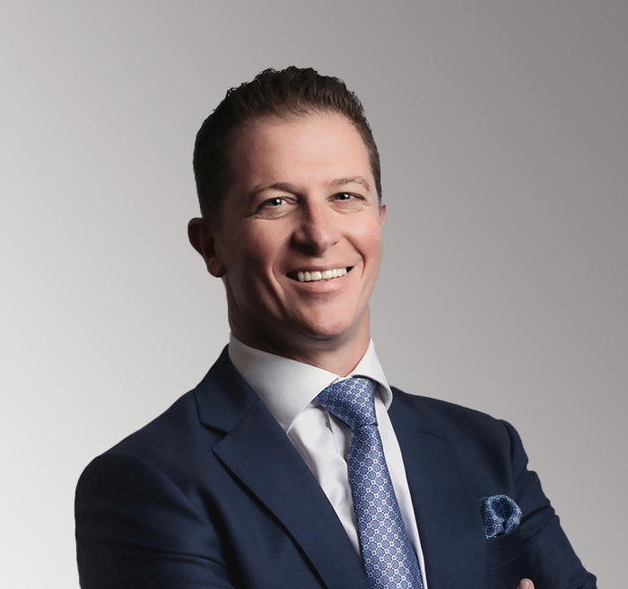 Smiling man in a navy suit, white shirt, and patterned blue tie posing confidently against a light grey gradient background.