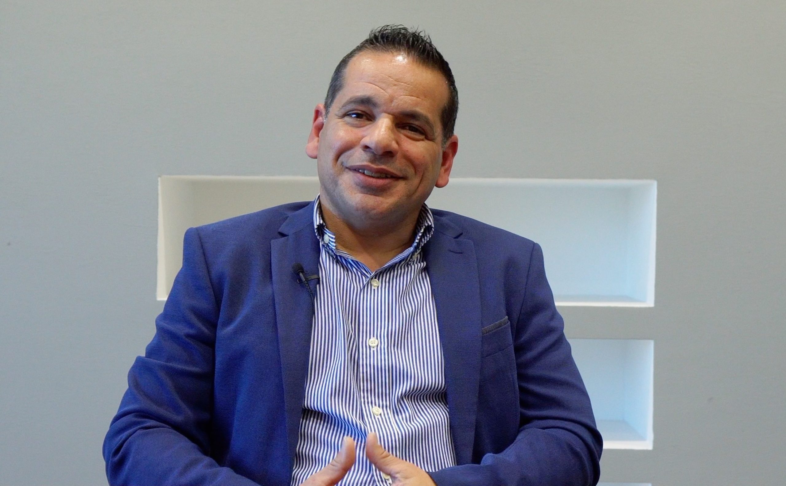 Smiling man wearing a blue blazer and striped shirt sitting against a light grey wall with a recessed shelf behind him.