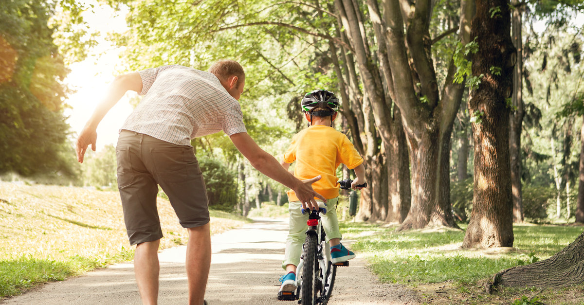 Adult helping a child ride a bicycle on a tree-lined path in a sunny park, symbolizing guidance, learning, and support.