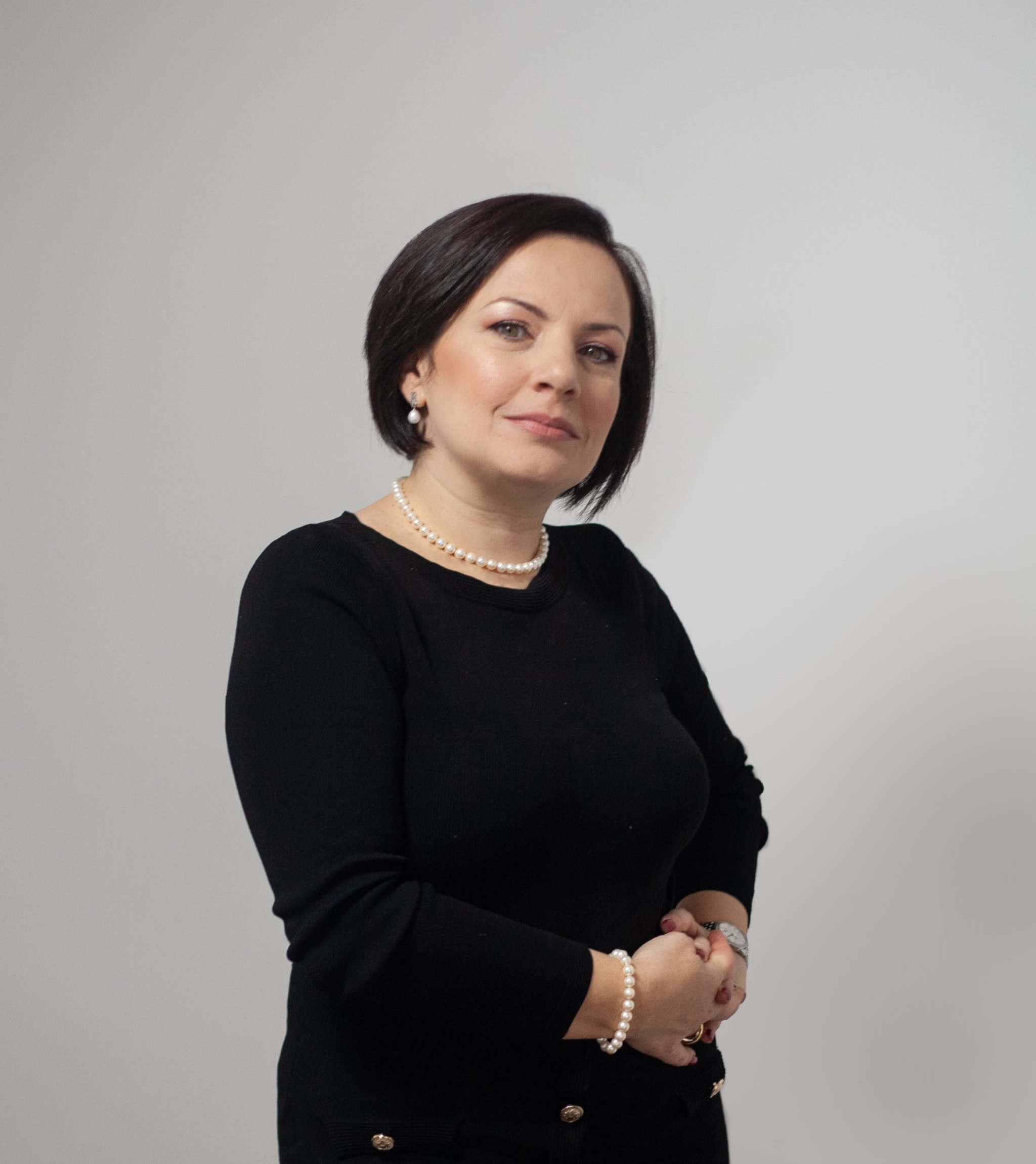 Woman with short dark hair wearing a black top and pearl jewelry, posing with hands clasped against a light grey background.