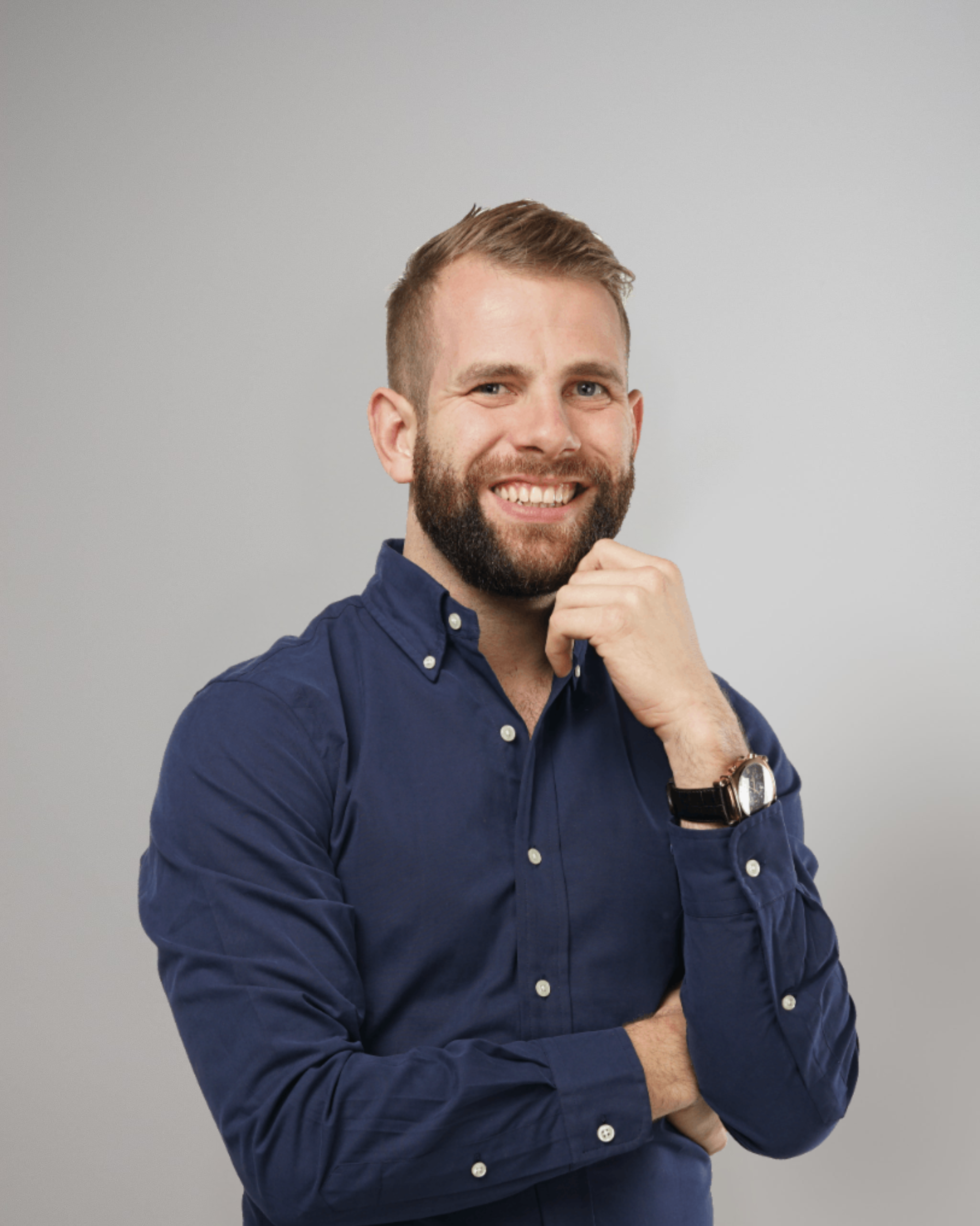 Smiling man with a beard wearing a navy blue button-up shirt, posing with one hand on his chin against a light grey background.