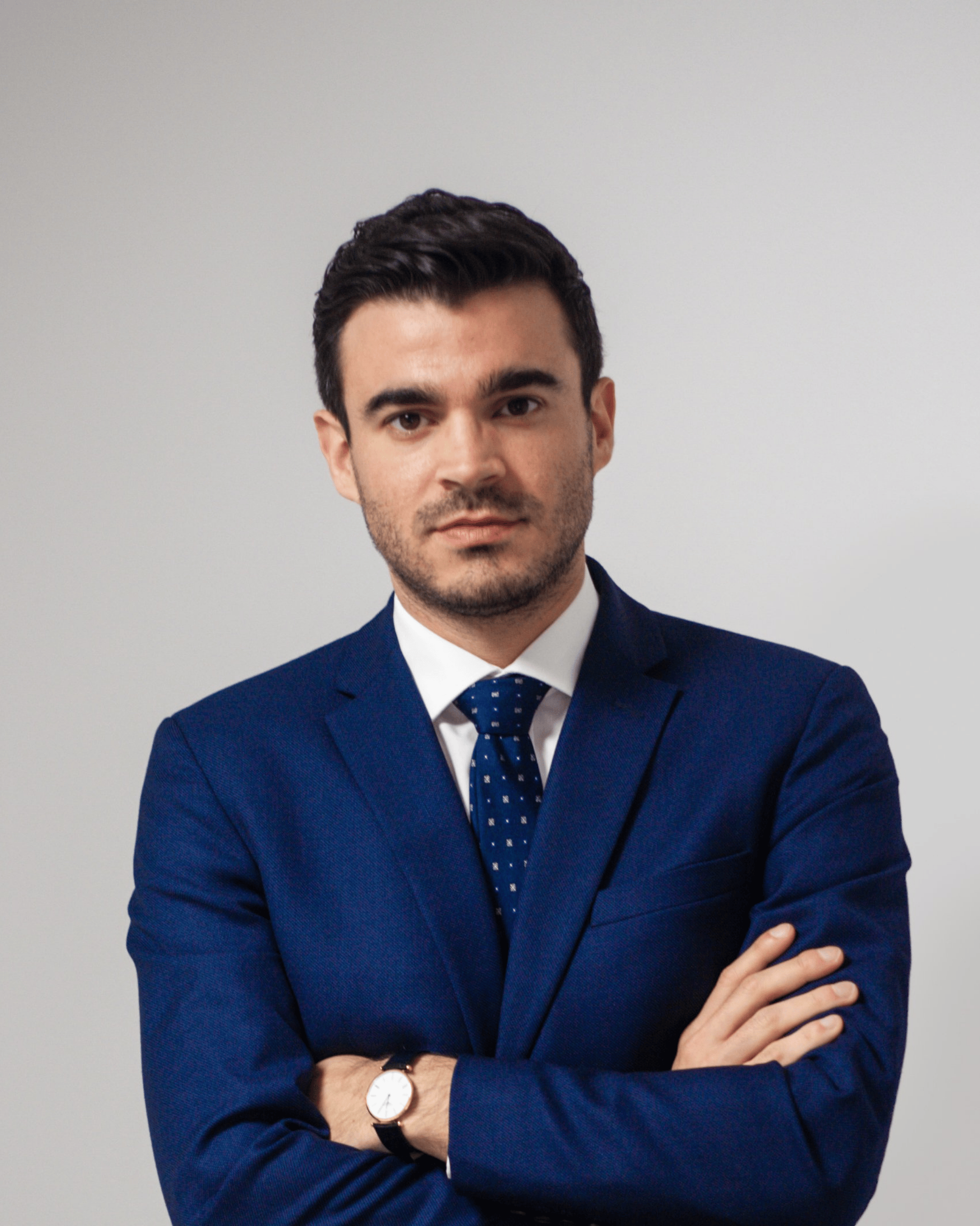 Man wearing a navy blue suit, white shirt, and dark blue patterned tie, standing with arms crossed against a light grey background.