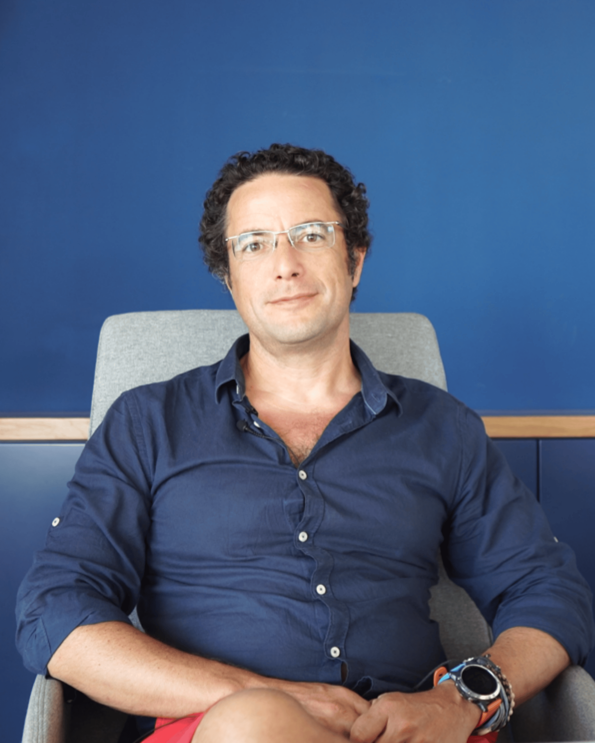 Man with curly hair and glasses sitting on a gray chair against a blue wall, wearing a navy blue shirt and red shorts.