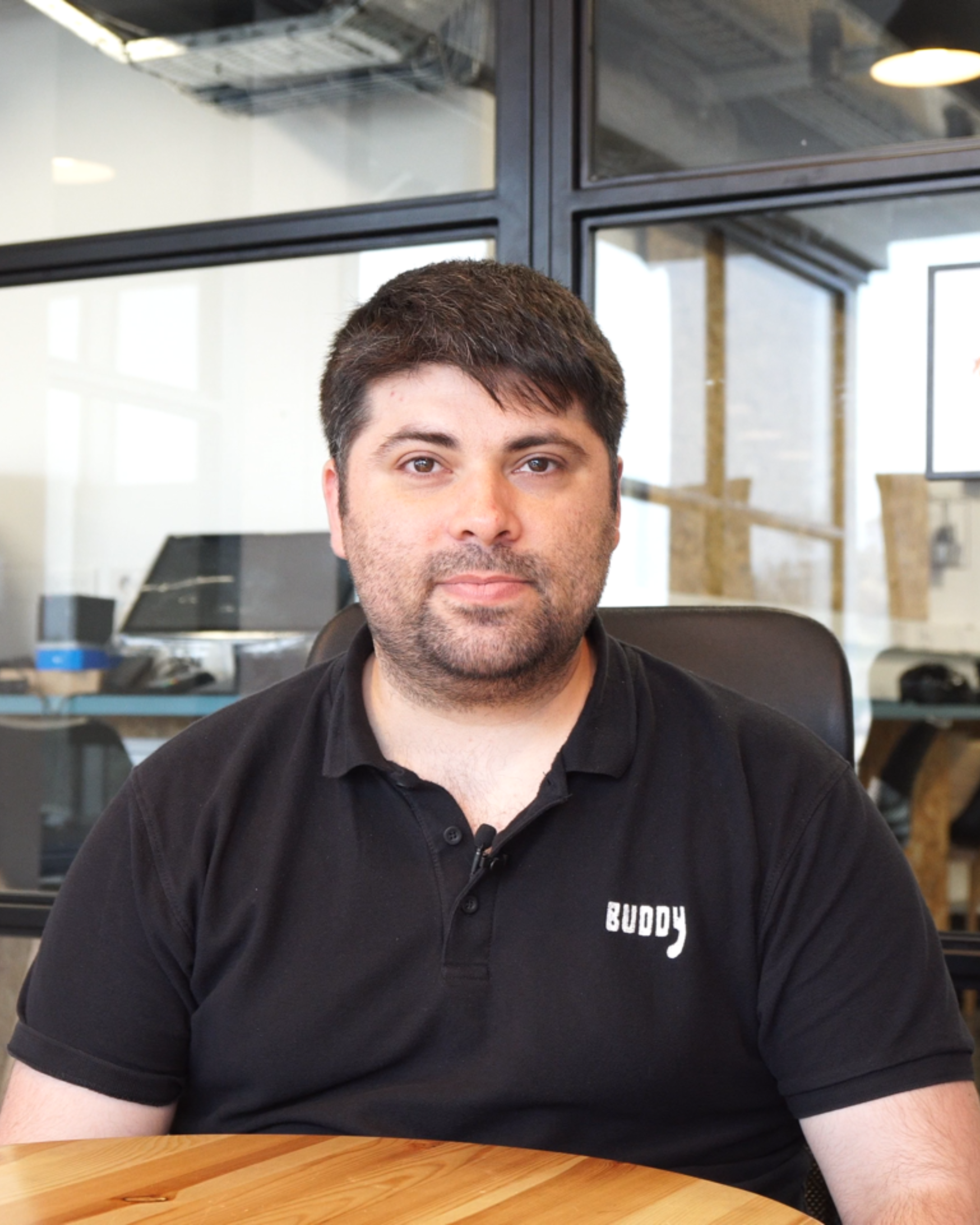 Man with short dark hair and beard wearing a black polo shirt with the word ‘BUDDY’ on it, seated at a wooden table in a modern office setting.