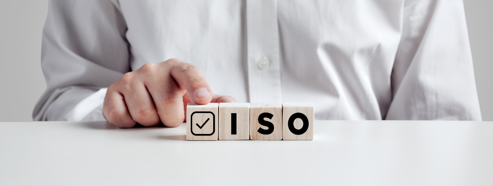 Person in a white shirt pointing at wooden blocks on a table that spell out ‘ISO’ with a checkmark symbol, representing ISO certification or quality standards.