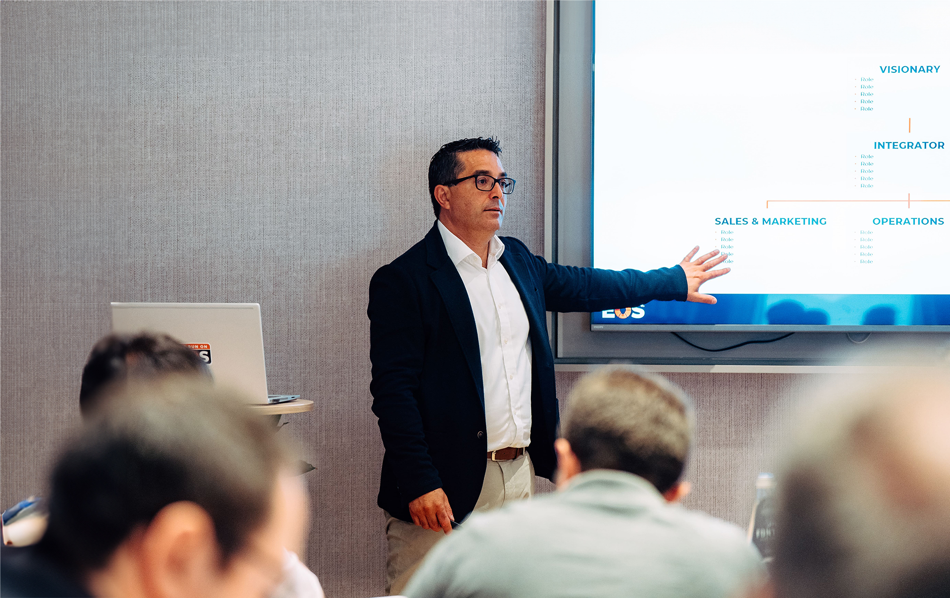 Man wearing glasses, a dark blazer, and a white shirt giving a presentation, pointing at a large screen displaying a business chart, with an audience seated in front of him.