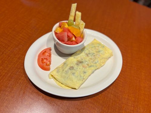 Plate with a folded omelette, a slice of tomato, and a cup of mixed fruit with grapes, watermelon, and orange segments on a wooden table.
