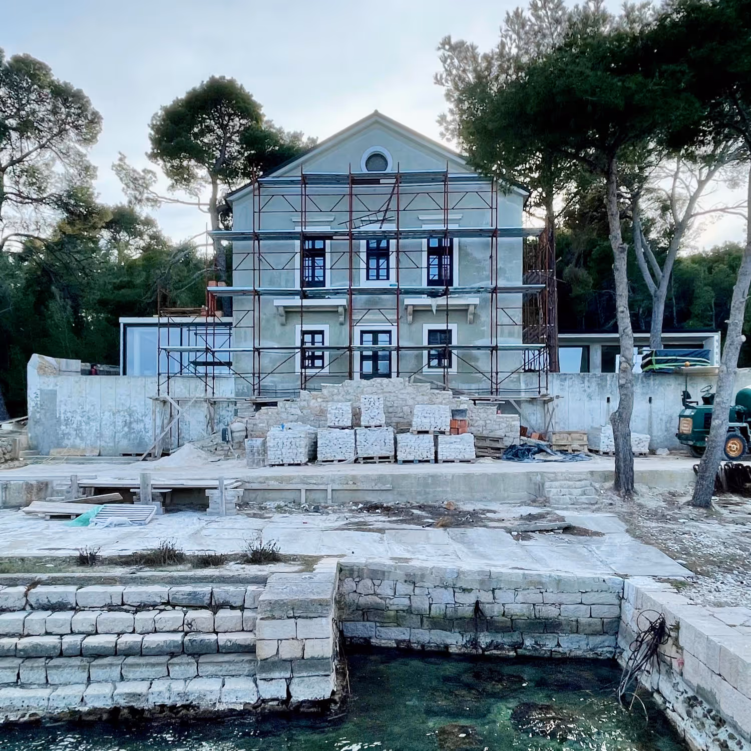 Services, build or renovate. Image showing a magnificent house in construction, with stone blocks and tiles ready to be laid in front of the façade, which has a scaffolding structure assembled in front. The house has a pine tree forest in the background and a private pier in the foreground.