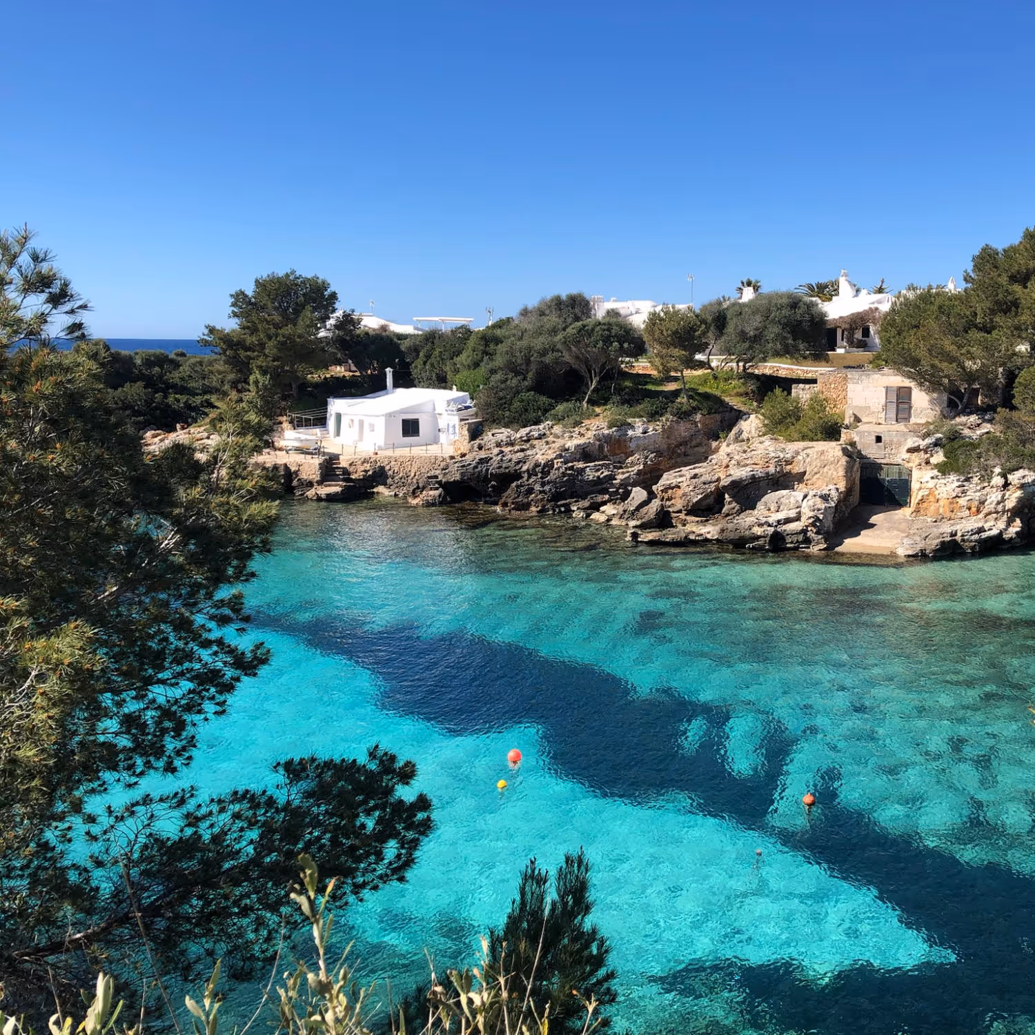 Image that shows Binisafuller cove and beach, with paradisiac turquoise calm waters. A white small house can be seen in the background, surrounded by nature.