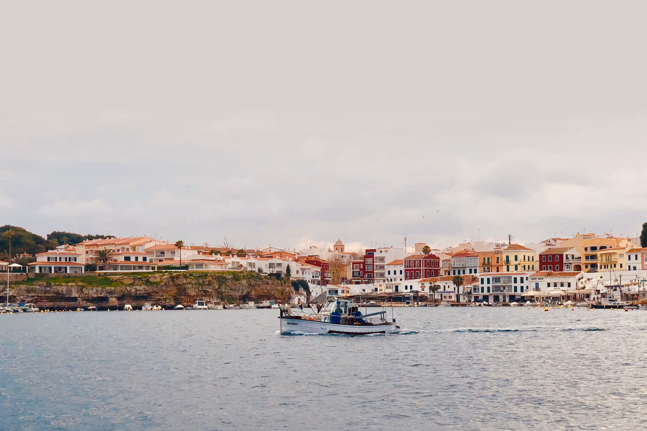 Contact. Image showing a traditional fishing llaut boat sailing through the Port of Mahon. Calesfonts can be seen in the background, with its typical and traditional red and yellow houses that crown the famous fishermen’s harbour in Es Castell.